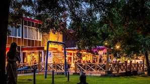 Un espace extérieur aménagé avec des tables et des chaises, une clôture en bois et, au fond, un bâtiment dont les fenêtres sont illuminées.