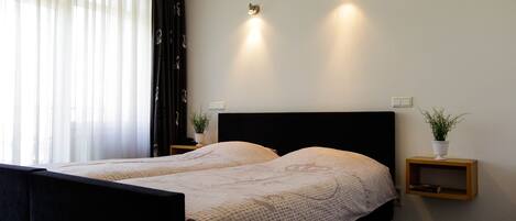 A double bed with a black headboard, two bedside tables with potted plants, and sheer curtains on a window.
