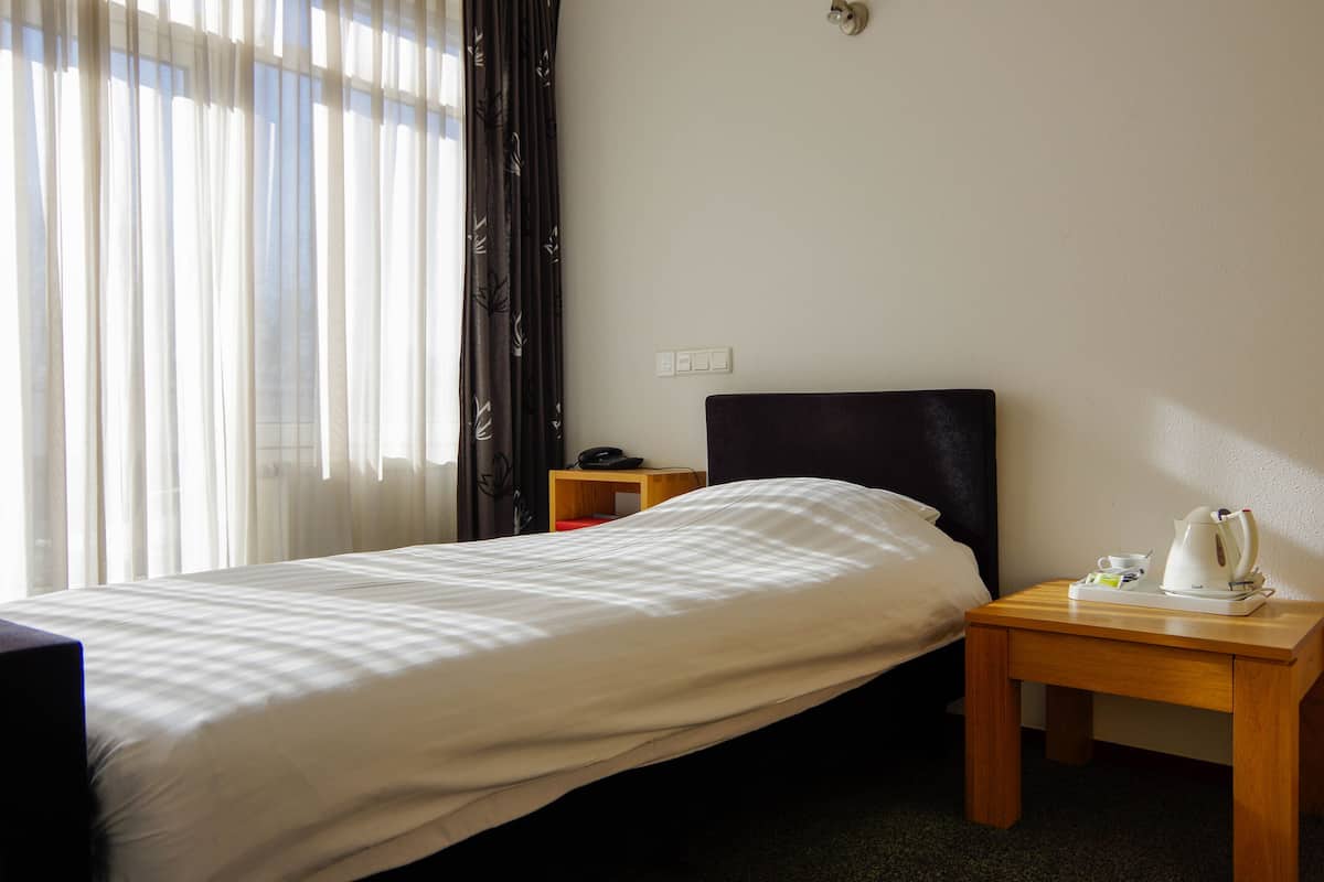 Een hotelkamer met een eenpersoonsbed, een nachtkastje met waterkoker, telefoon en een klein plankje.