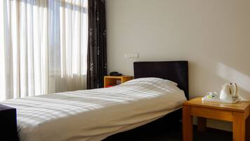 Habitación de hotel con una cama individual, una mesita de noche con tetera, teléfono y un estante pequeño.