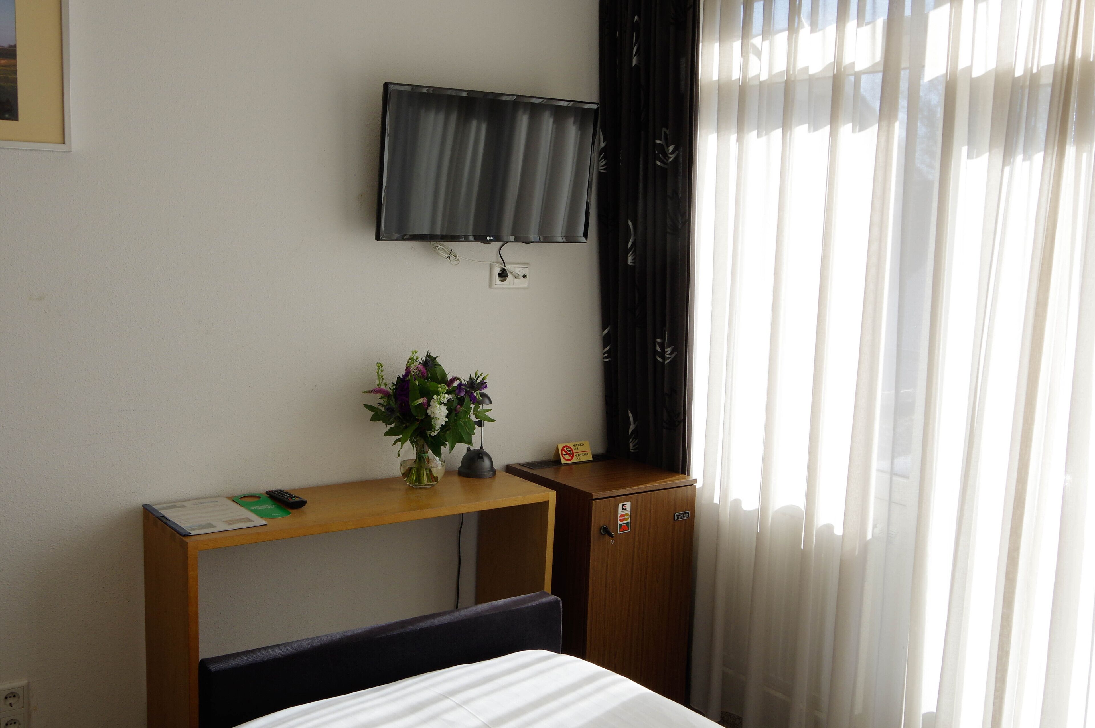 A hotel room with a bed, a desk with a vase of flowers, a television mounted on the wall, and a wooden cabinet.