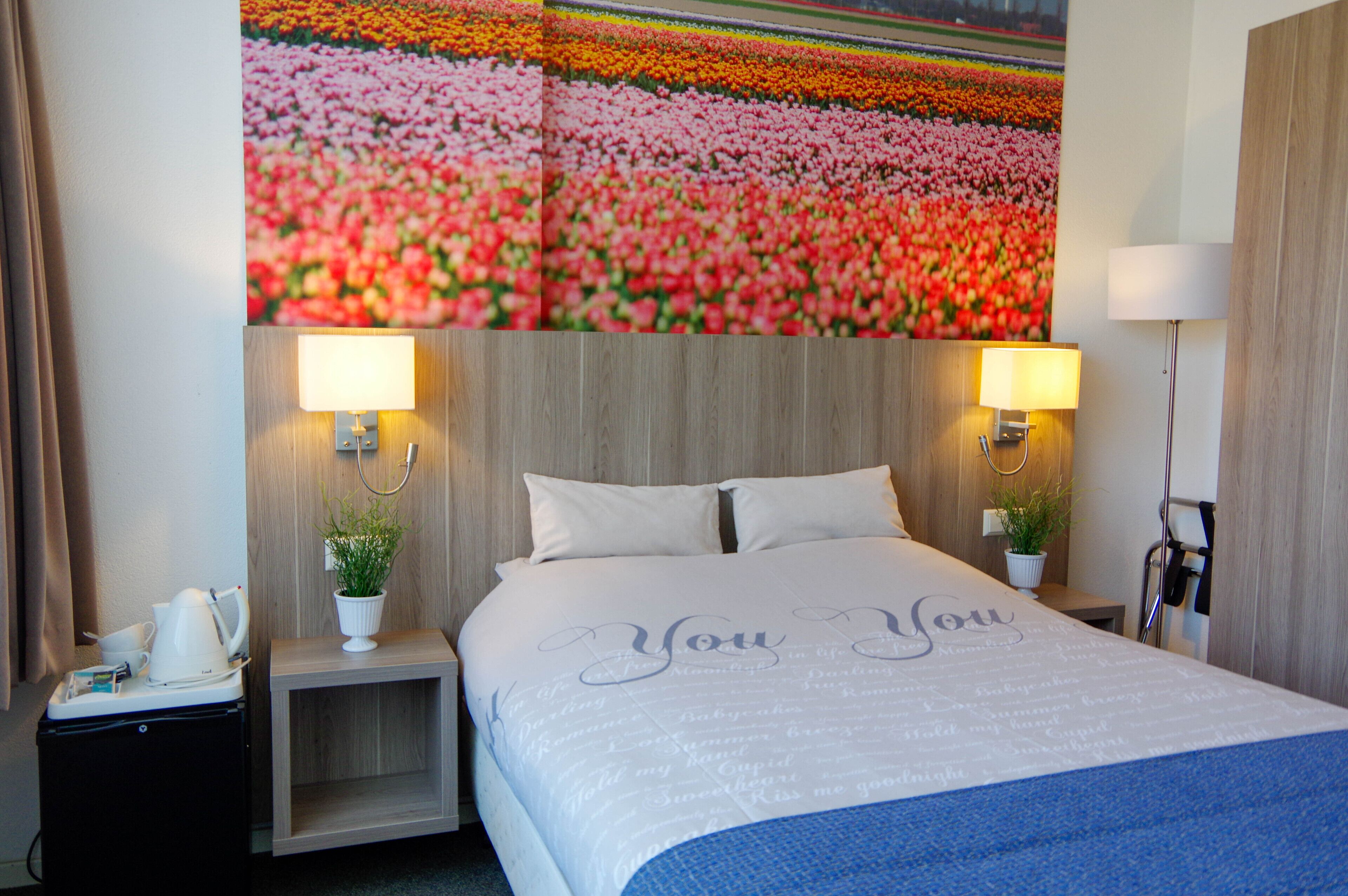 A hotel room with a large floral mural on the wall, a bed with a 'You & You' bedspread, two bedside lamps, a small bedside table with a plant, and a kettle.