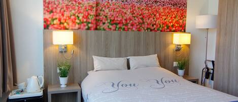 Quarto de hotel com um grande mural floral na parede, uma cama com colcha 'You & You', duas luminárias de cabeceira, uma mesinha de cabeceira com uma planta e uma chaleira.