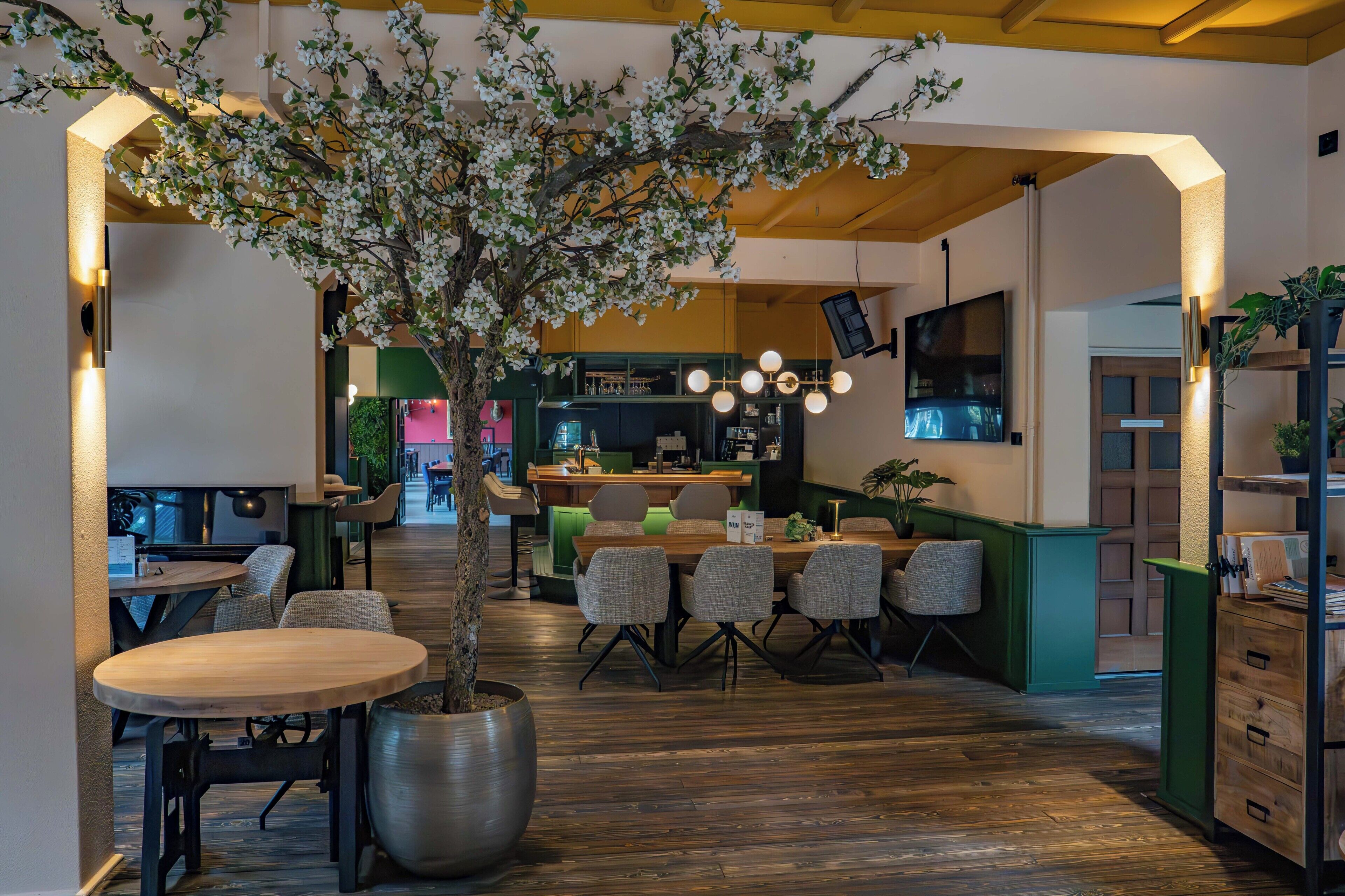 A restaurant interior with a central dining area, wooden flooring, and a large potted tree.