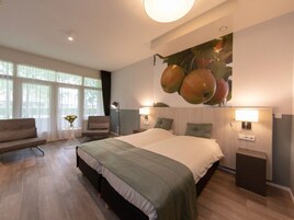 Una habitación de hotel moderna con una cama grande, una zona de estar con sofá y sillones, una mesita con un jarrón y una lámpara fijada a la pared.