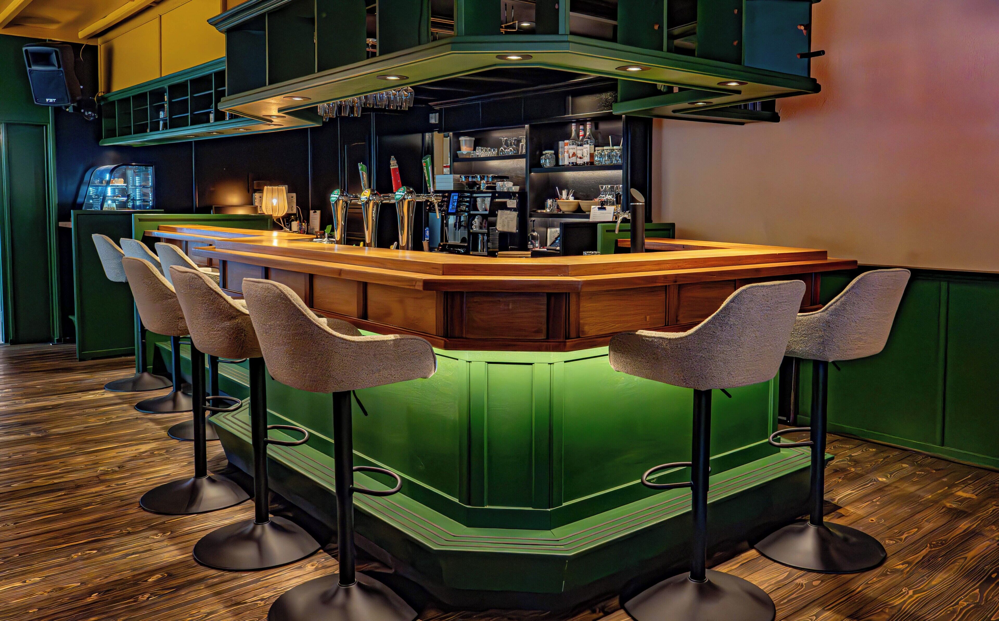 Zona de bar con mostrador verde, taburetes y una variedad de botellas y vasos.