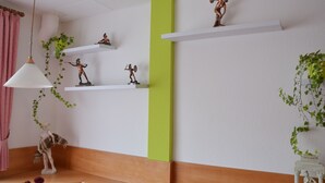 Een kamer met witte wanden, een groene accentmuur en houten lambrisering onderaan. Er zijn drie witte planken met beelden, een hangende plant en een witte lamp.