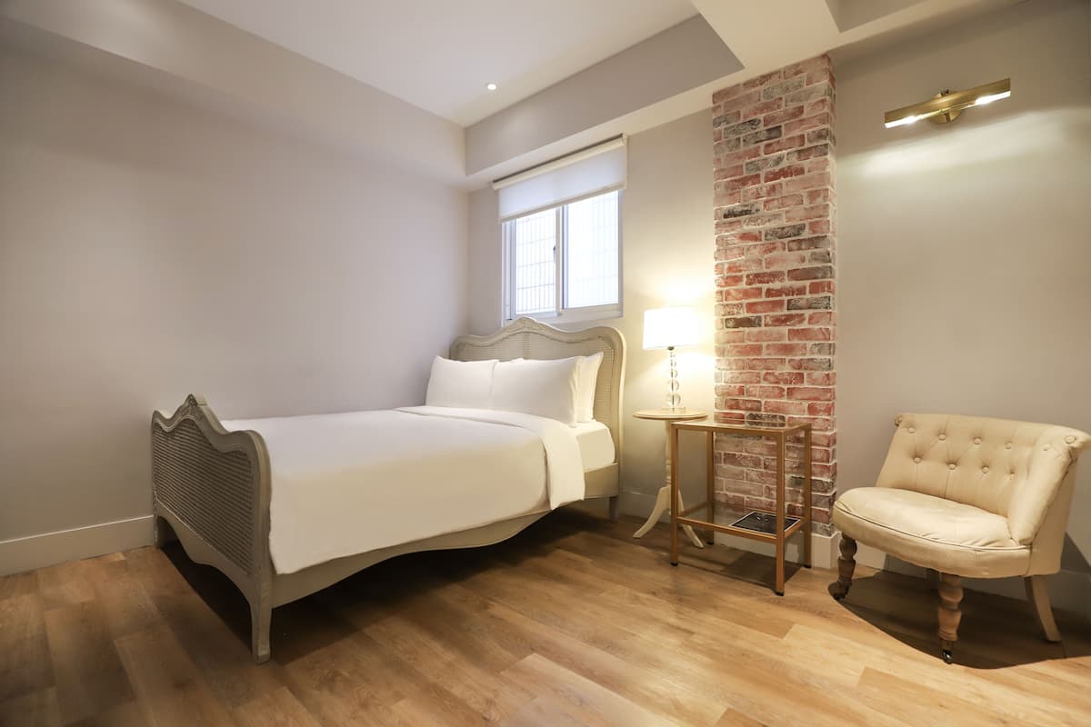 A bedroom with a bed, a chair, a lamp, and a brick wall.