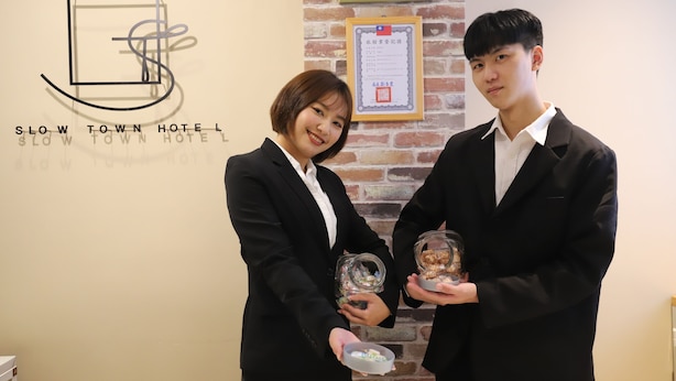 Two individuals holding awards in front of a wall with a hotel sign and a certificate. - Slow Town Hotel - Reel (Taipei)