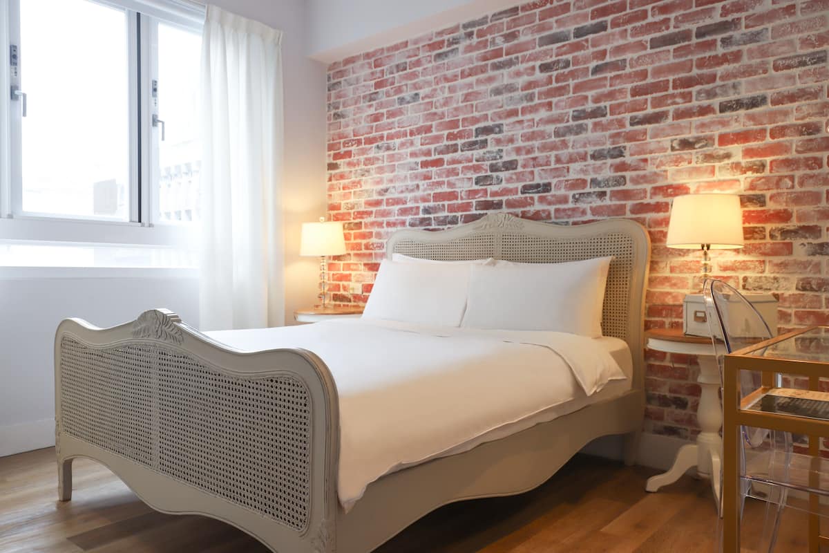 A bedroom with a brick wall, a bed with white linens, two bedside lamps, and a window with curtains.