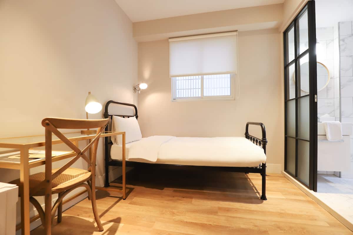 A small, well-lit room with a bed, a desk, and a chair. There is a window with blinds, a wall-mounted light, and a view of a bathroom through a glass door.