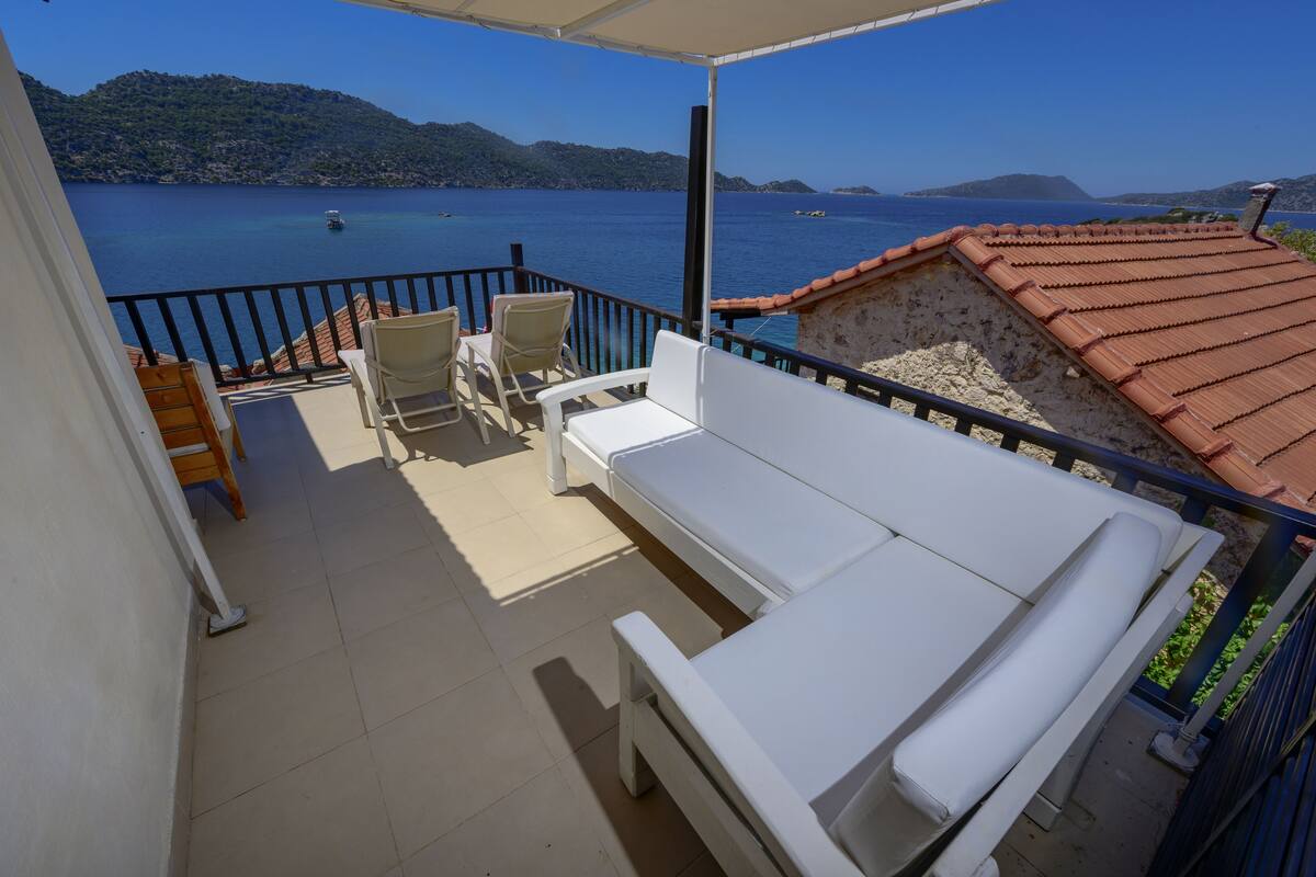 A balcony with a white sofa, two chairs, and a small table. The balcony overlooks a body of water with boats and distant hills.