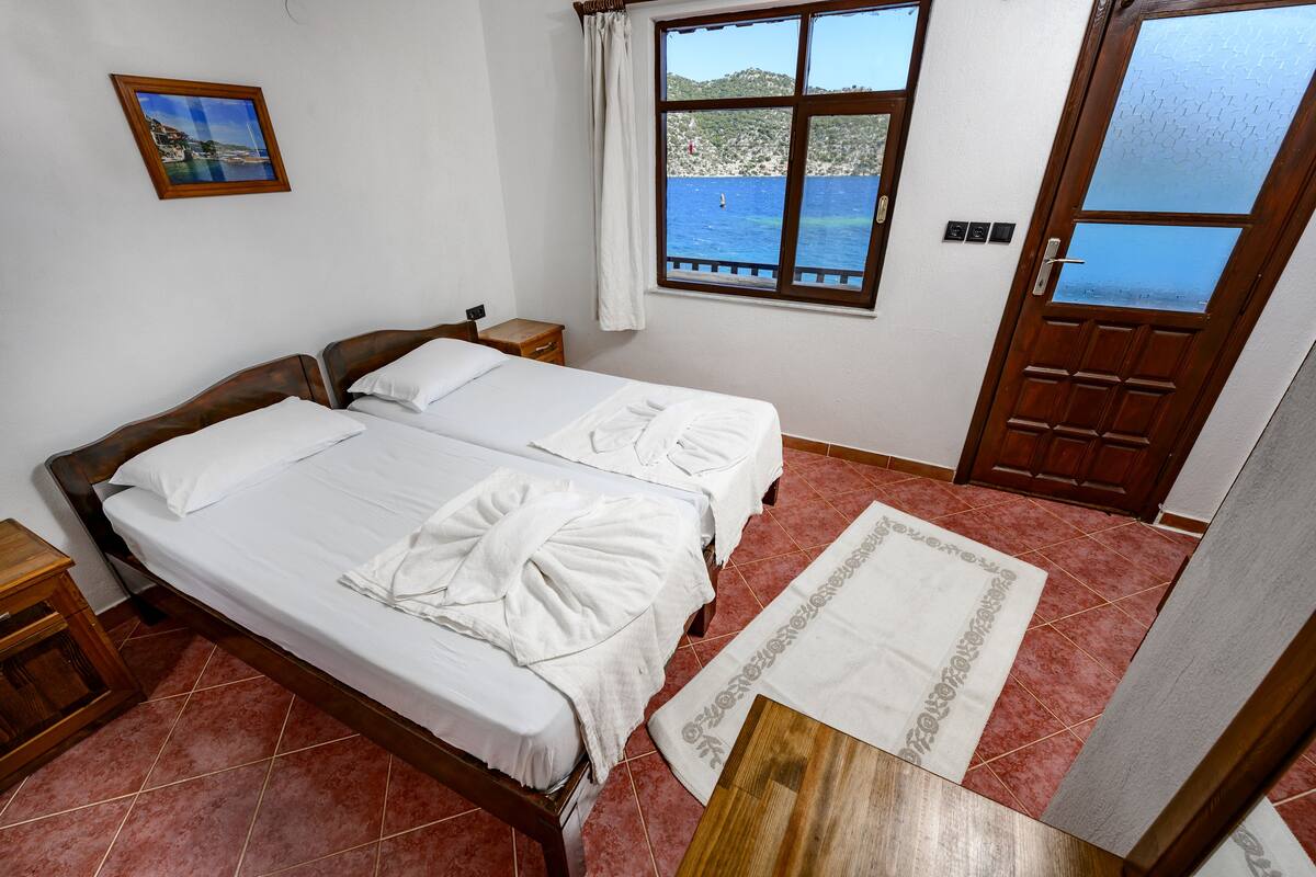 A double bed with white linens and a wooden headboard, a small bedside table, a window with a view of the sea, and a wooden door.