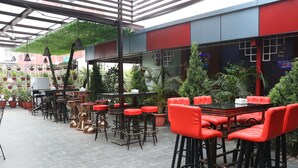 Un café al aire libre con taburetes y mesas rojos, rodeado de plantas en macetas y un techo verde.