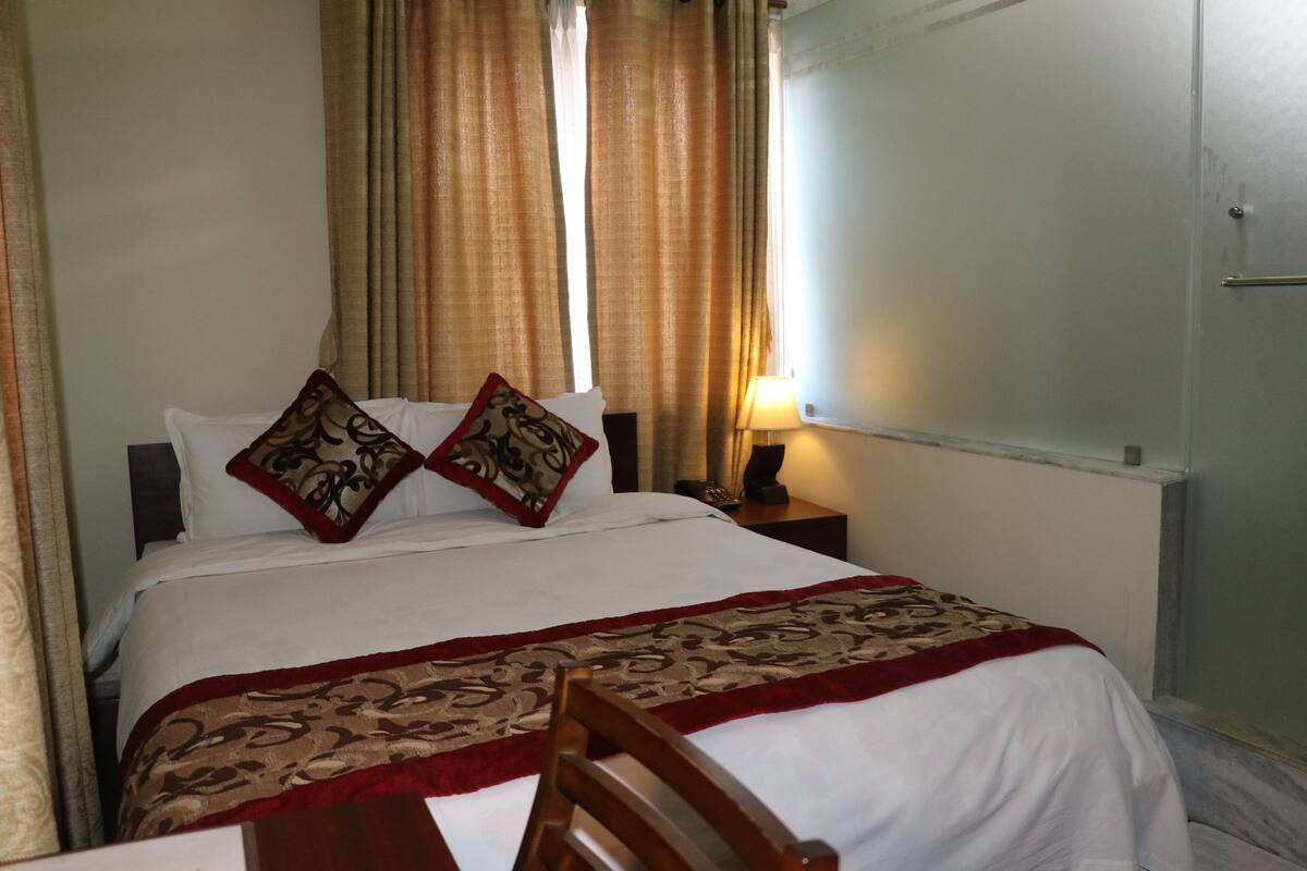 A neatly made bed with a patterned bedspread and pillows, a bedside table with a lamp, and a chair in front of the bed.