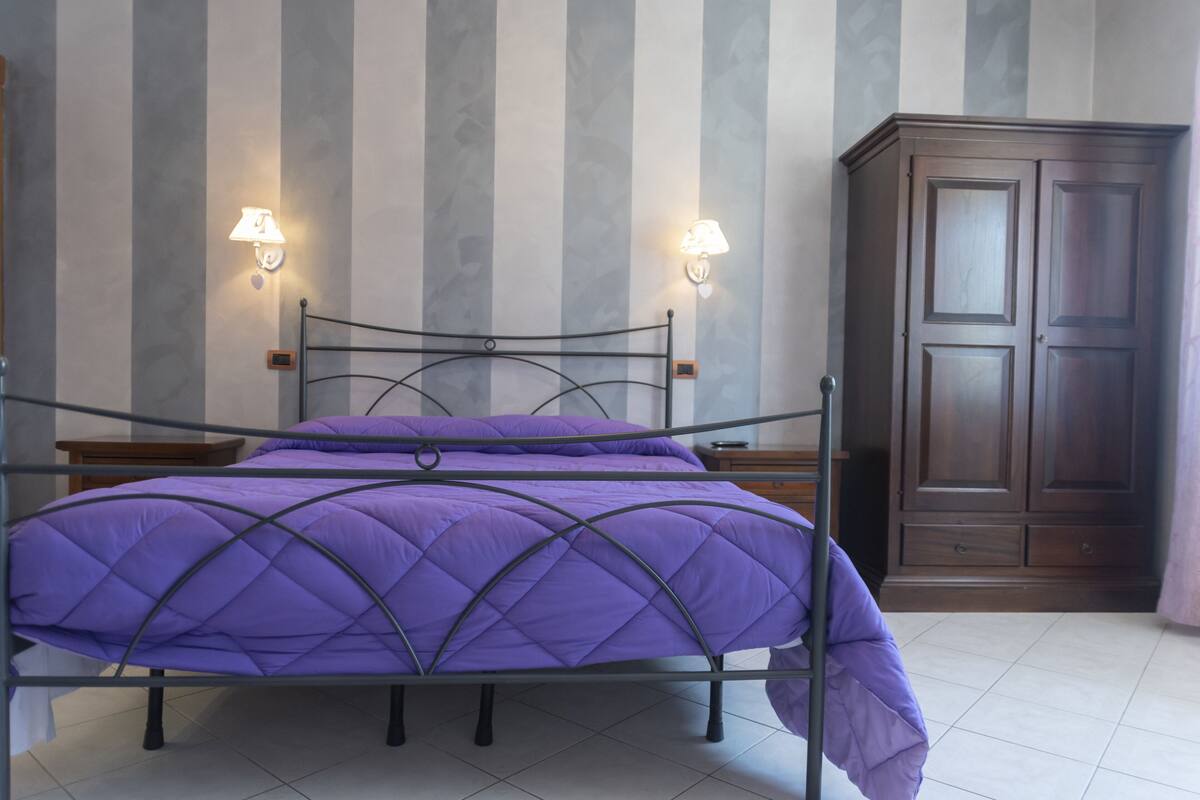 Un lit avec un cadre en métal et une literie violette, deux lampes fixées au mur et une armoire en bois.