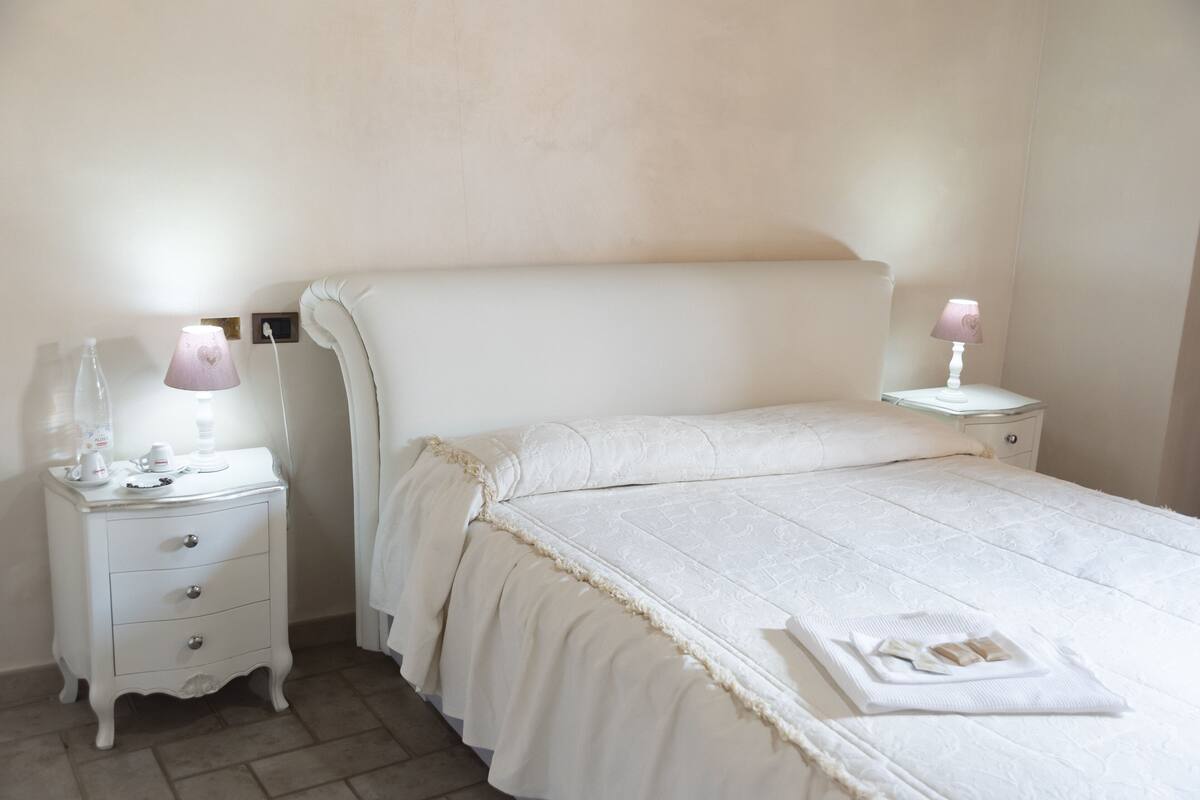 Un lit bien fait, avec une couette blanche et des oreillers, une table de chevet avec une lampe et une petite commode.