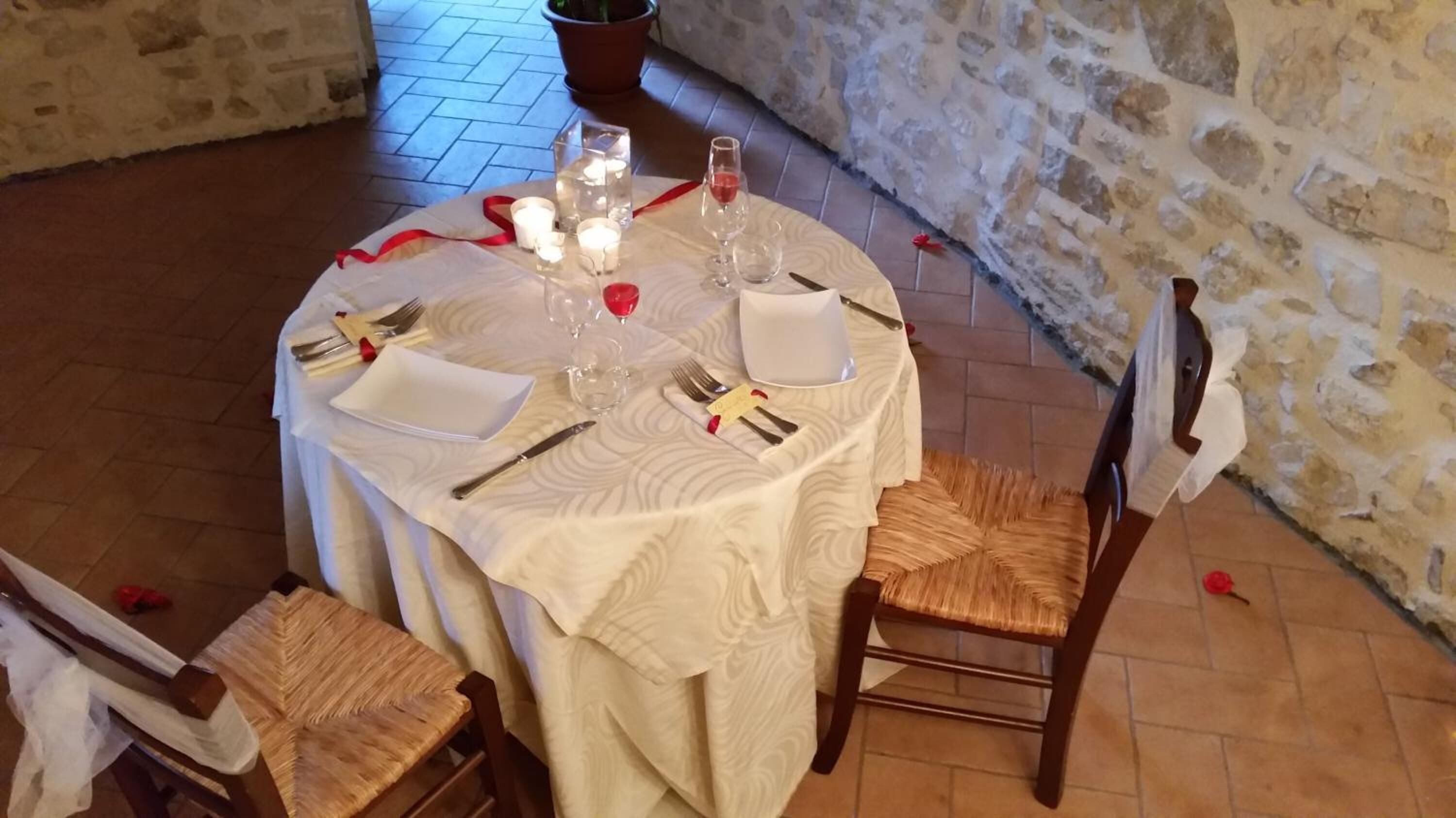 A round dining table with a white tablecloth, set with plates, cutlery, glasses, and candles. There are two wooden chairs with woven seats. The room has stone walls and a tiled floor.