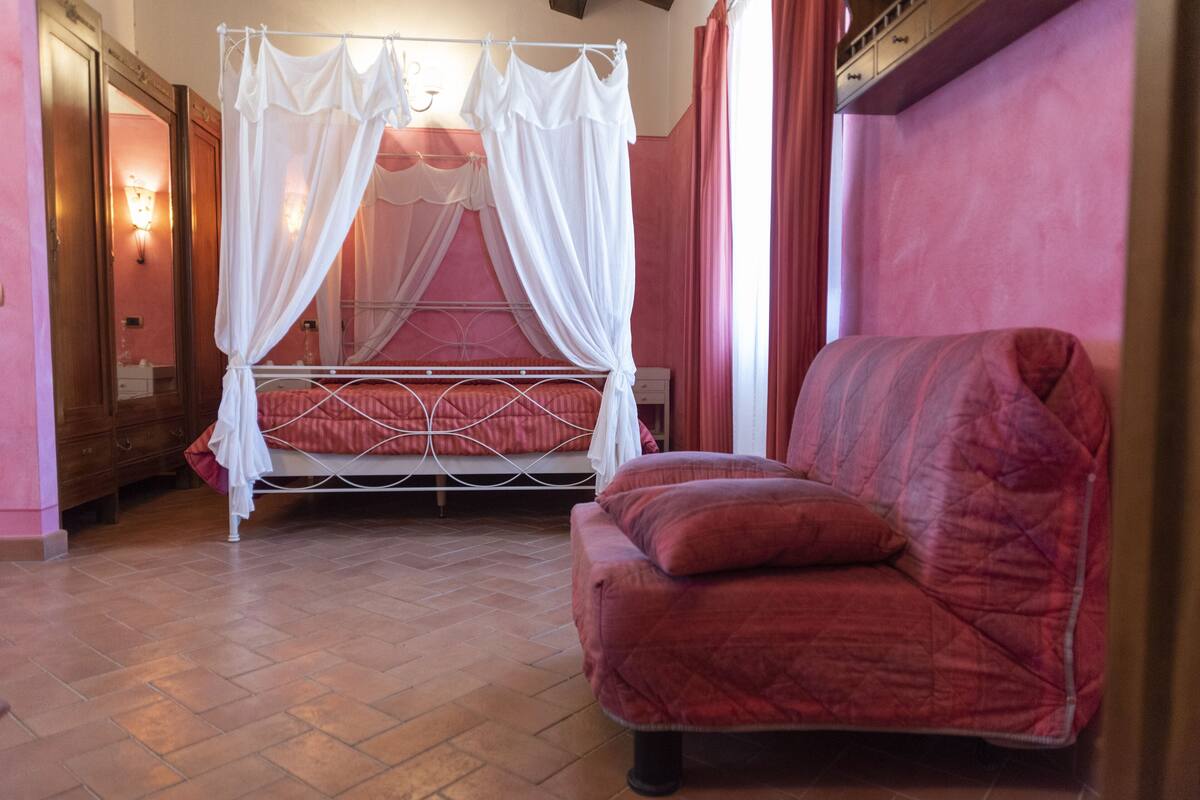Une chambre à coucher avec un lit à baldaquin, un fauteuil rouge recouvert de tissu et un parquet à motifs chevrons.