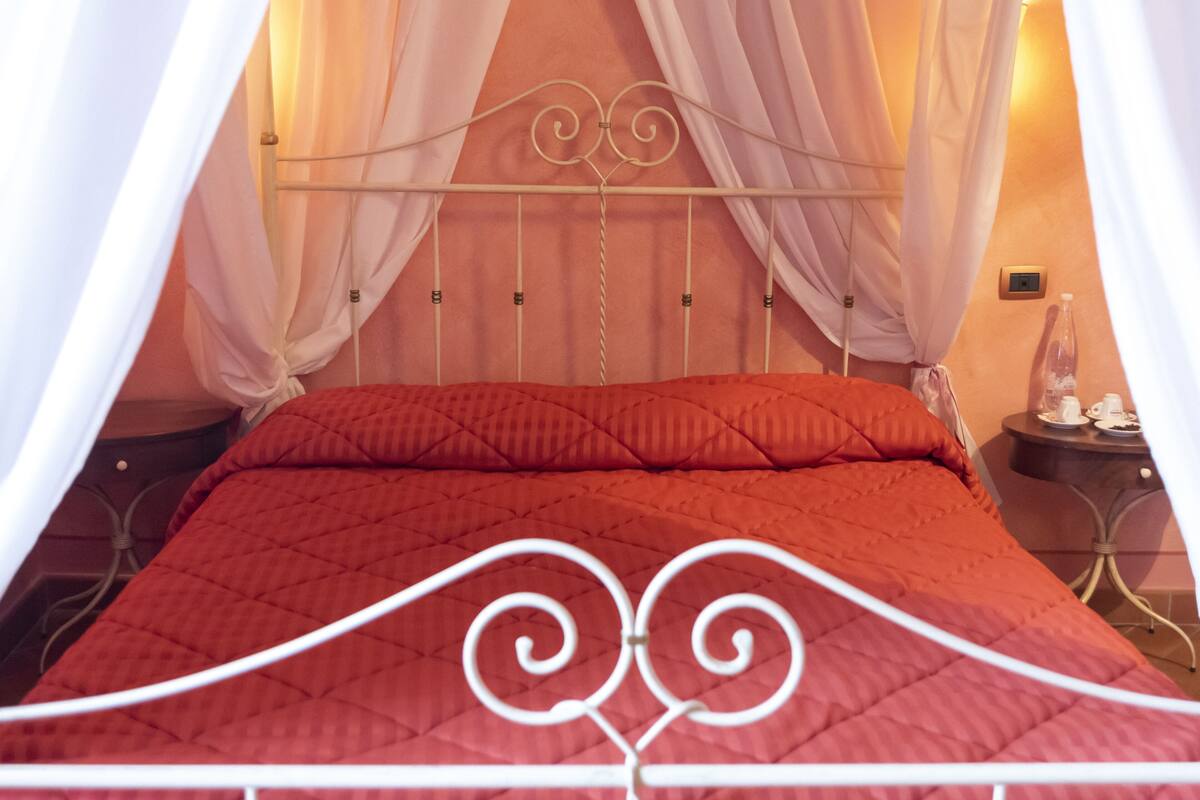 Un lit avec une couette rouge, des rideaux blancs et une tête de lit en métal.