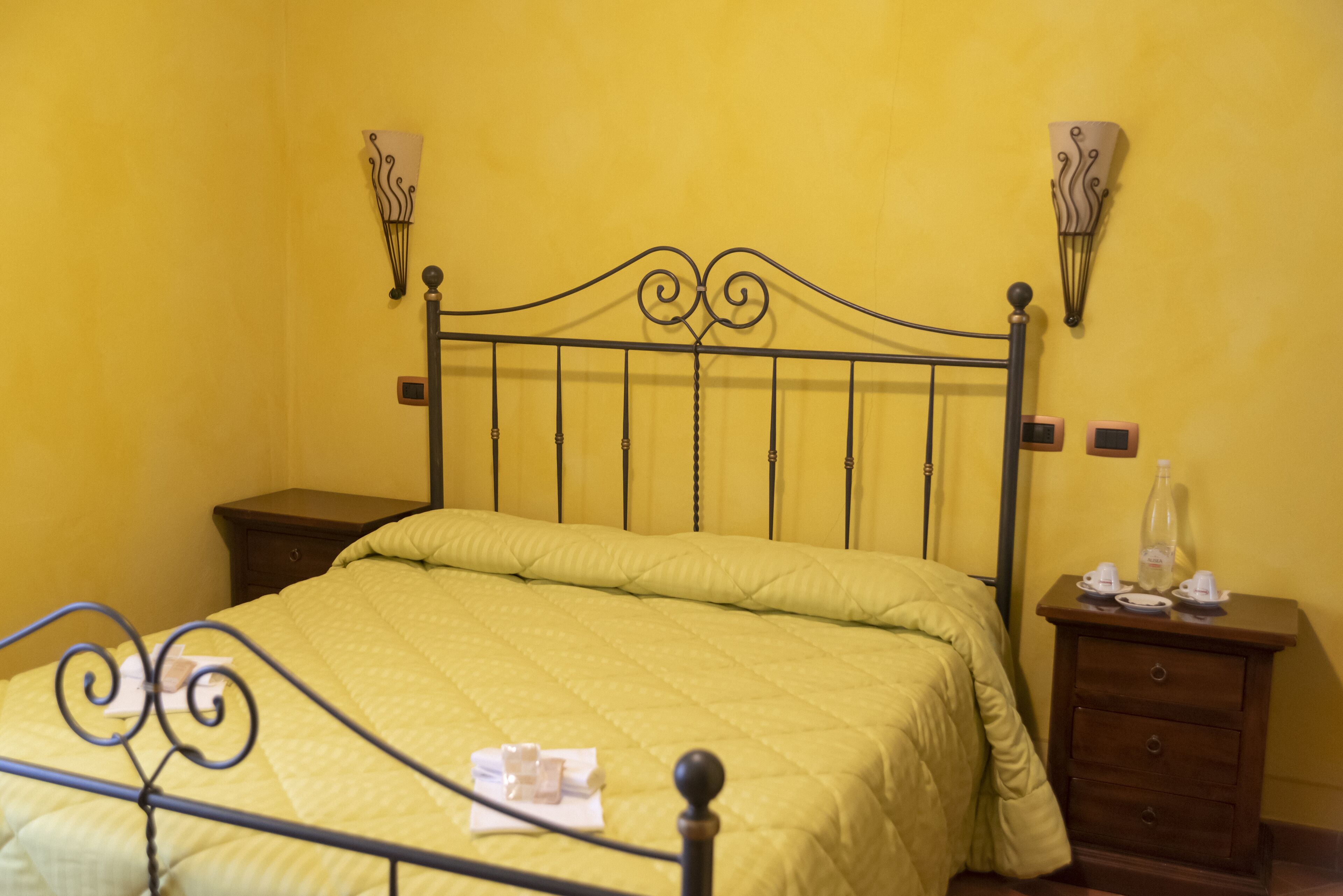 A bed with a metal frame, a yellow wall, and a nightstand with a bottle and glasses.