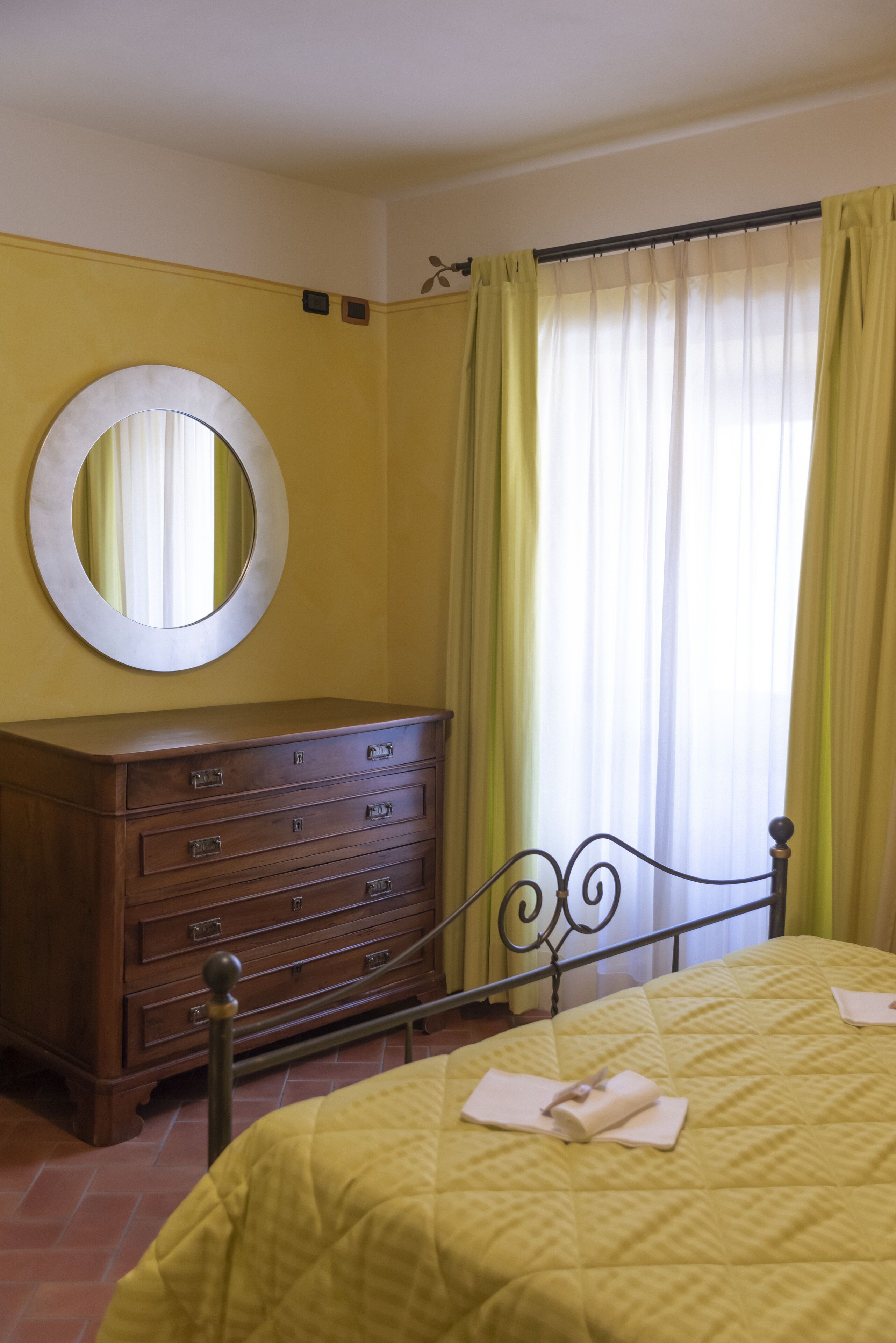 Een kamer met een bed, een houten dressoir, een ronde spiegel en een raam met gele gordijnen.