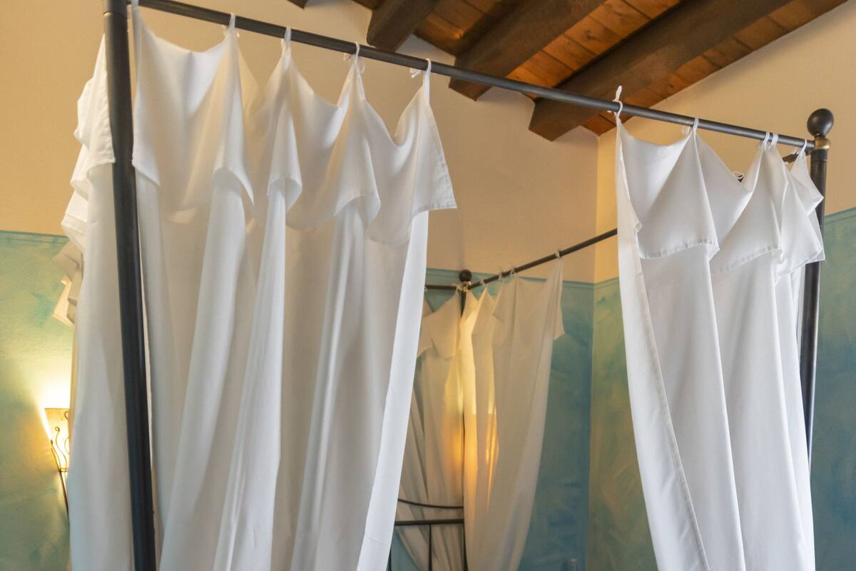 Un lit avec une couette bleue et des rideaux blancs, une table de chevet avec une bouteille, et un mur avec un luminaire.