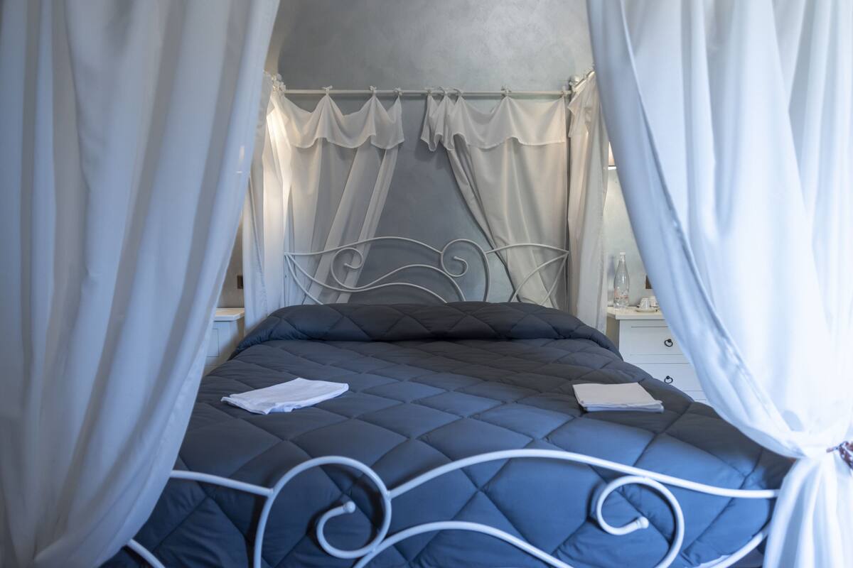 Un lit avec un matelas matelassé, des rideaux blancs et une tête de lit en métal.