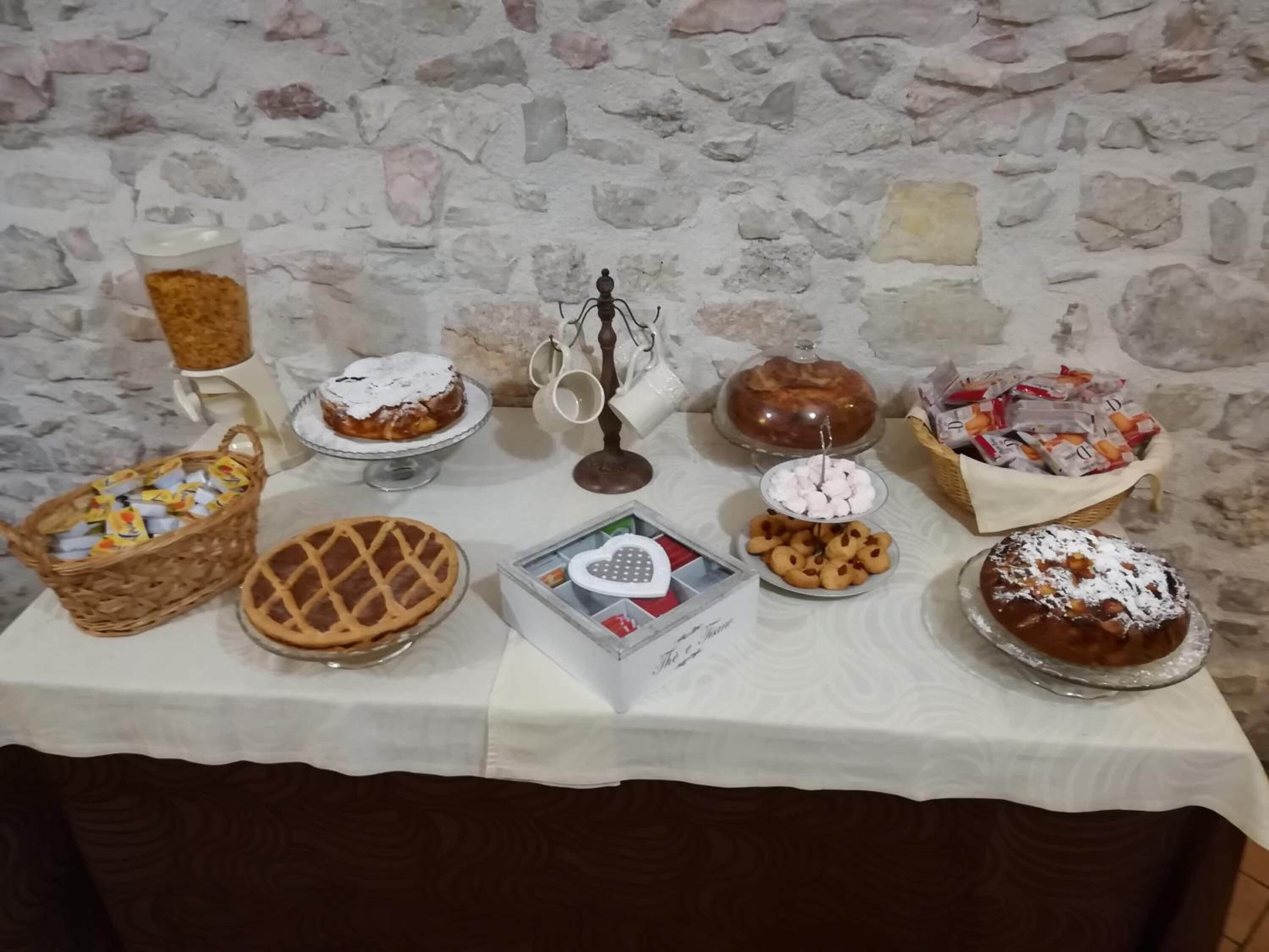 Een tafel met diverse desserts en snacks, waaronder taarten, koekjes en een gebak, voor een achtergrond van een stenen muur.