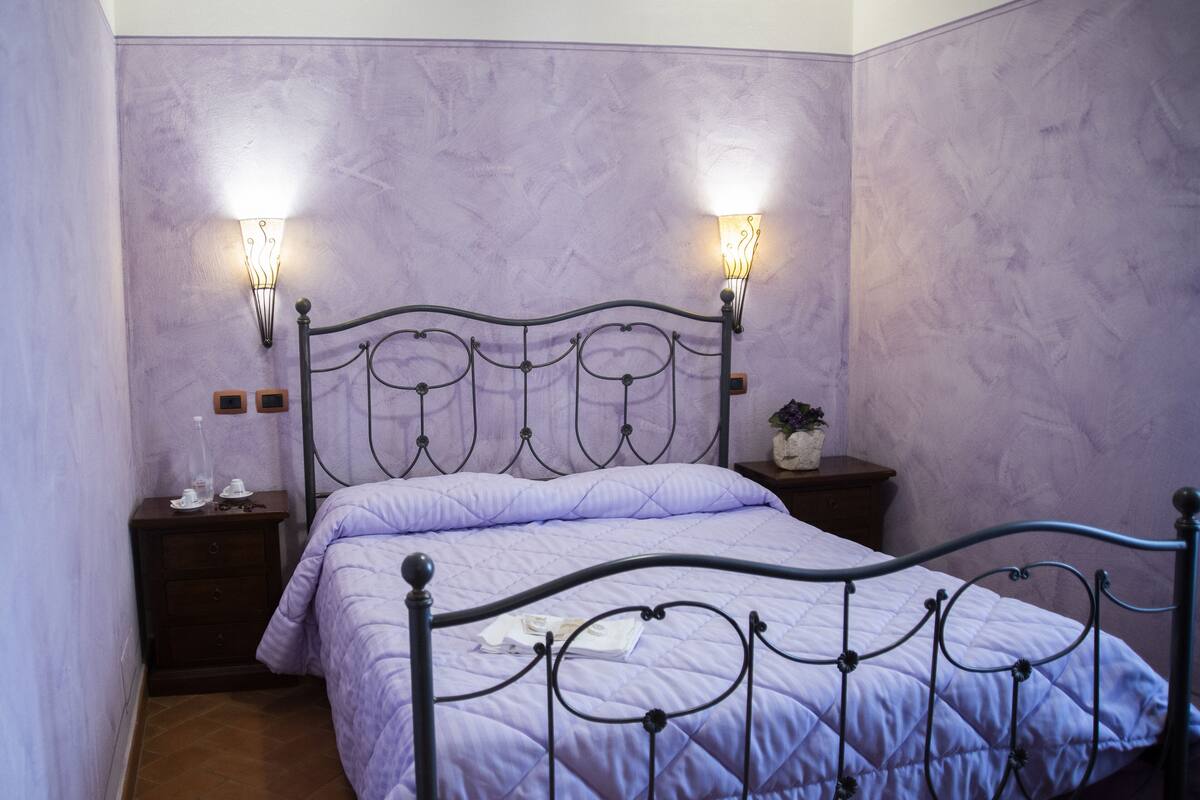 Un lit avec un cadre en métal et une couvre-lit matelassée de couleur violette, deux lampes fixées au mur, une petite table de chevet avec une plante, et une table de nuit avec une bouteille et des verres.