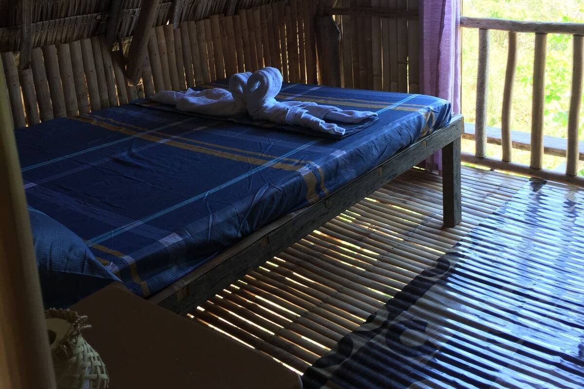 A room with a bed, a table, and a hanging bag. The room has a thatched roof and wooden flooring.