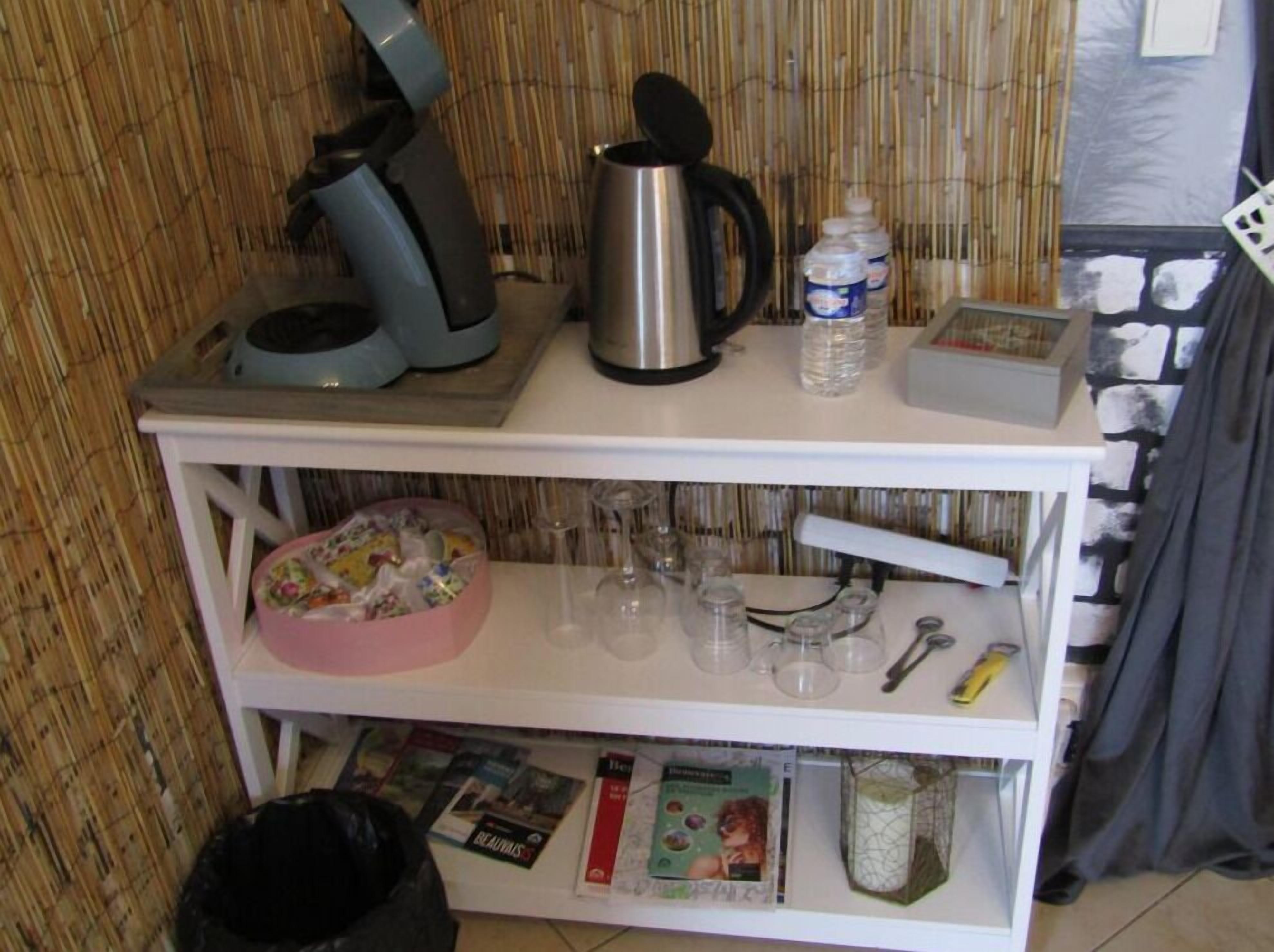 A kitchenette with a coffee maker, kettle, and water dispenser on the countertop. Below, there are shelves with a pink dish rack, glasses, and a small basket.