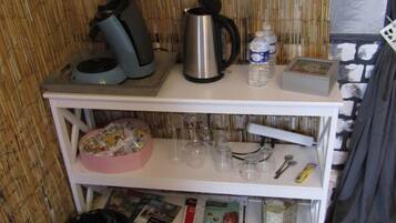 A kitchenette with a coffee maker, kettle, and water dispenser on the countertop. Below, there are shelves with a pink dish rack, glasses, and a small basket.