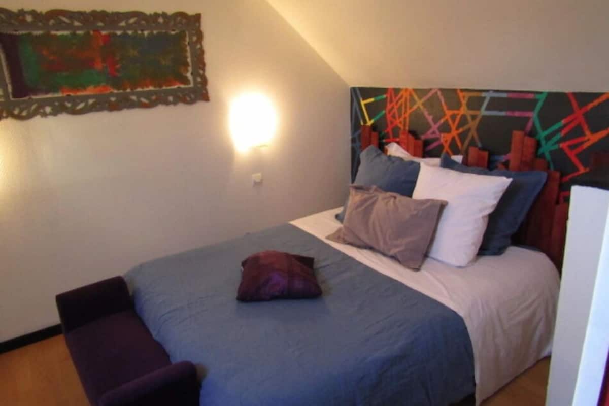 Une chambre à coucher avec un lit, des oreillers, un banc et une peinture abstraite aux couleurs vives accrochée au mur.
