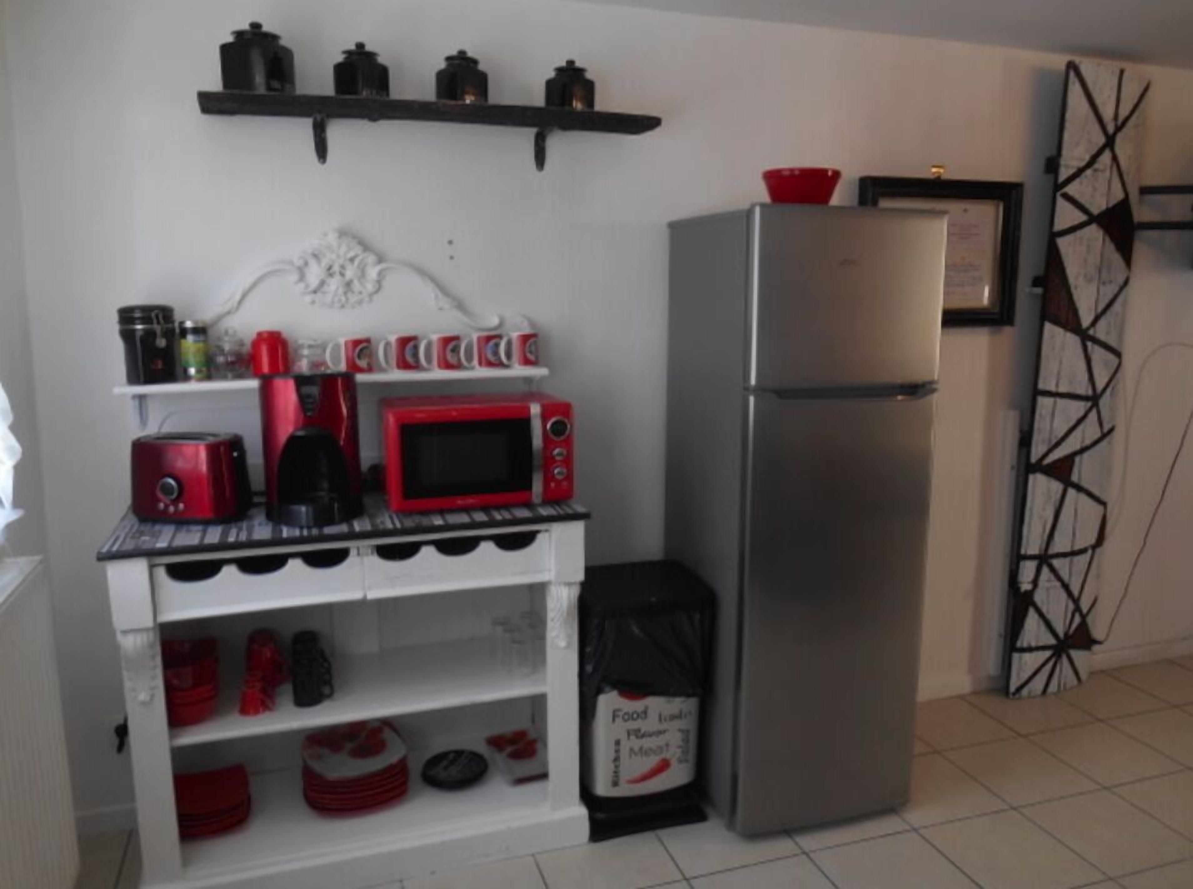 A compact kitchen with a microwave, toaster, and coffee maker on a white counter. A stainless steel refrigerator is to the right, and a trash bin is in front.