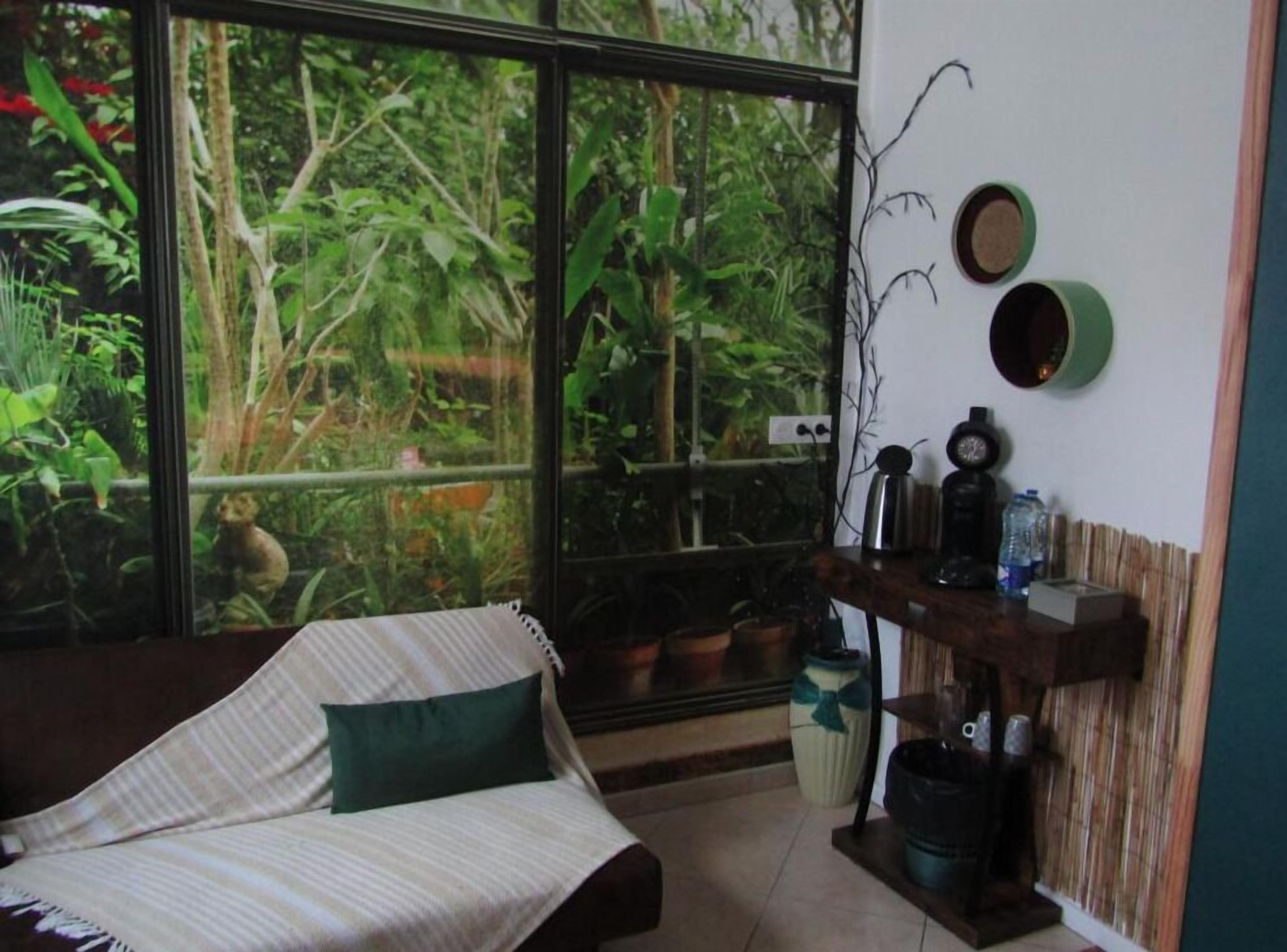 Una habitación con un ventanal grande que da a un jardín exuberante, un sofá a rayas con un cojín verde, y una estantería con varios objetos, incluyendo un reloj y una botella de agua.