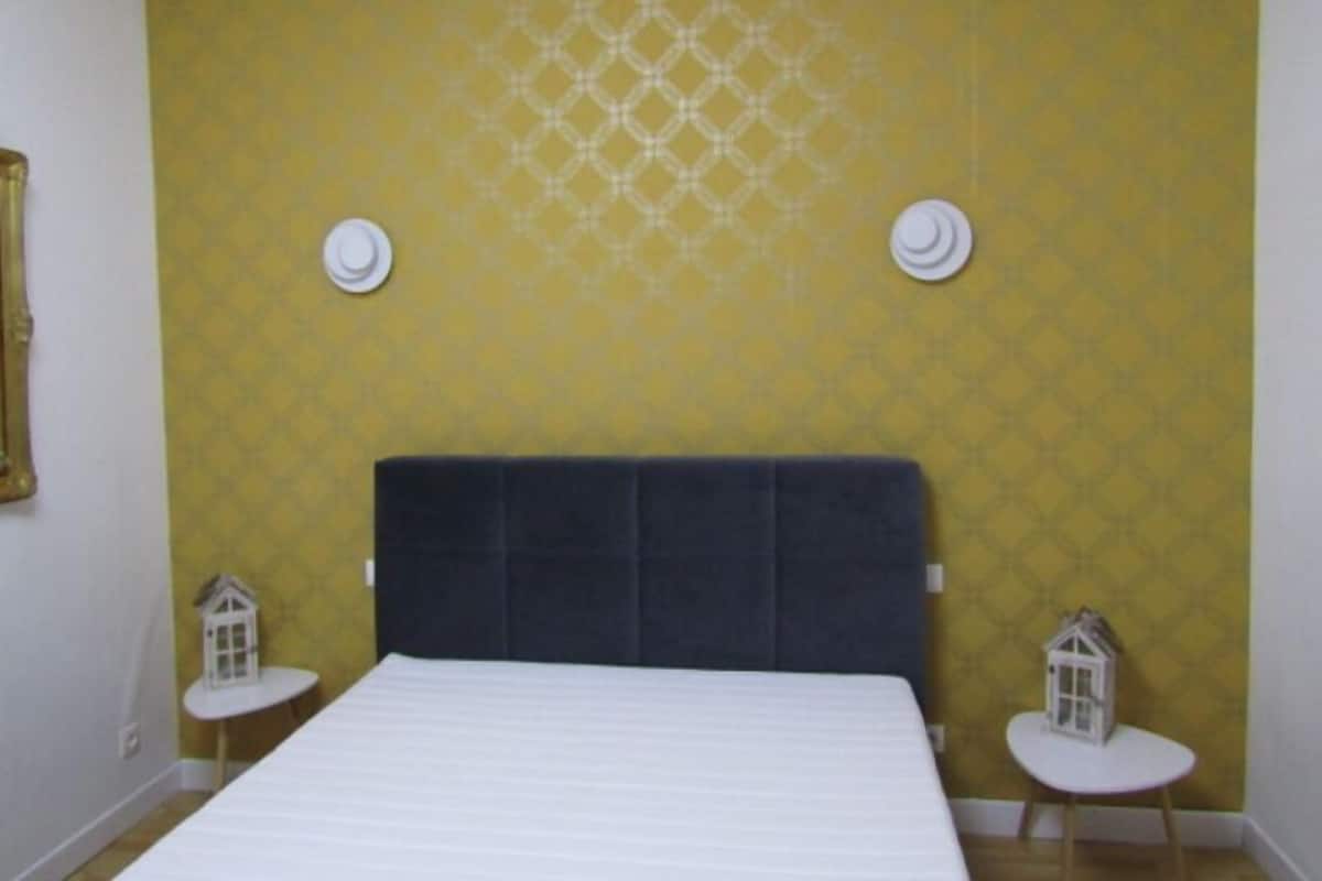 Une chambre avec un grand lit, un papier peint jaune à motifs, deux petites tables blanches décorées de motifs représentant des maisons, et une tête de lit sombre.