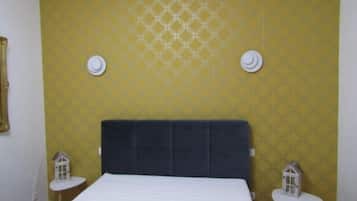 A bedroom with a large bed, yellow patterned wallpaper, two small white tables with house-shaped decorations, and a dark headboard.