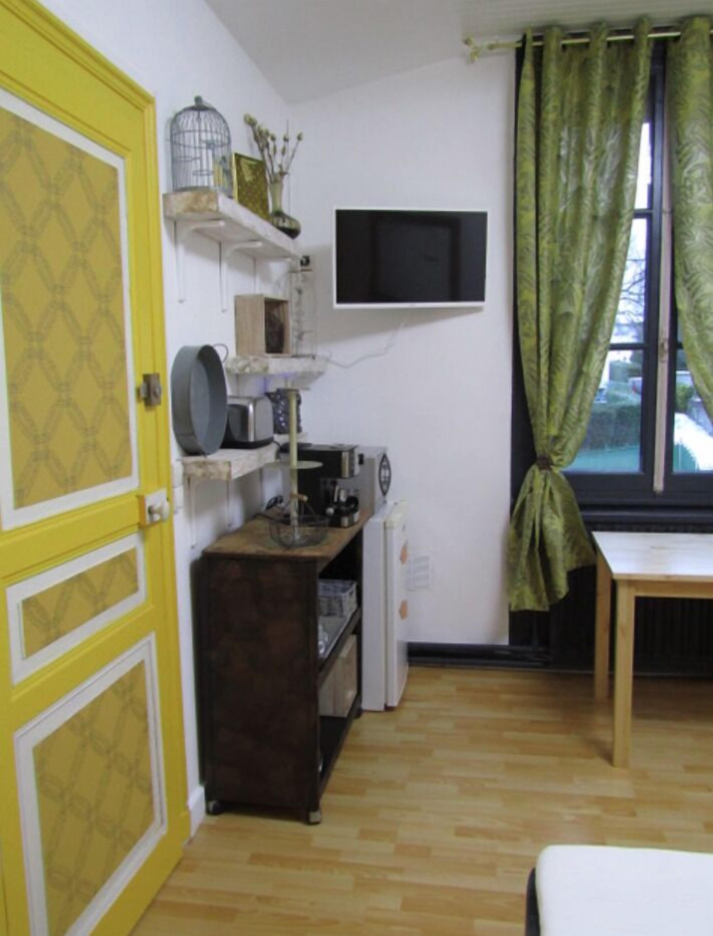 A compact kitchen with a microwave, kettle, and a small refrigerator. A yellow door with a geometric pattern is visible, and a window with green curtains is present.
