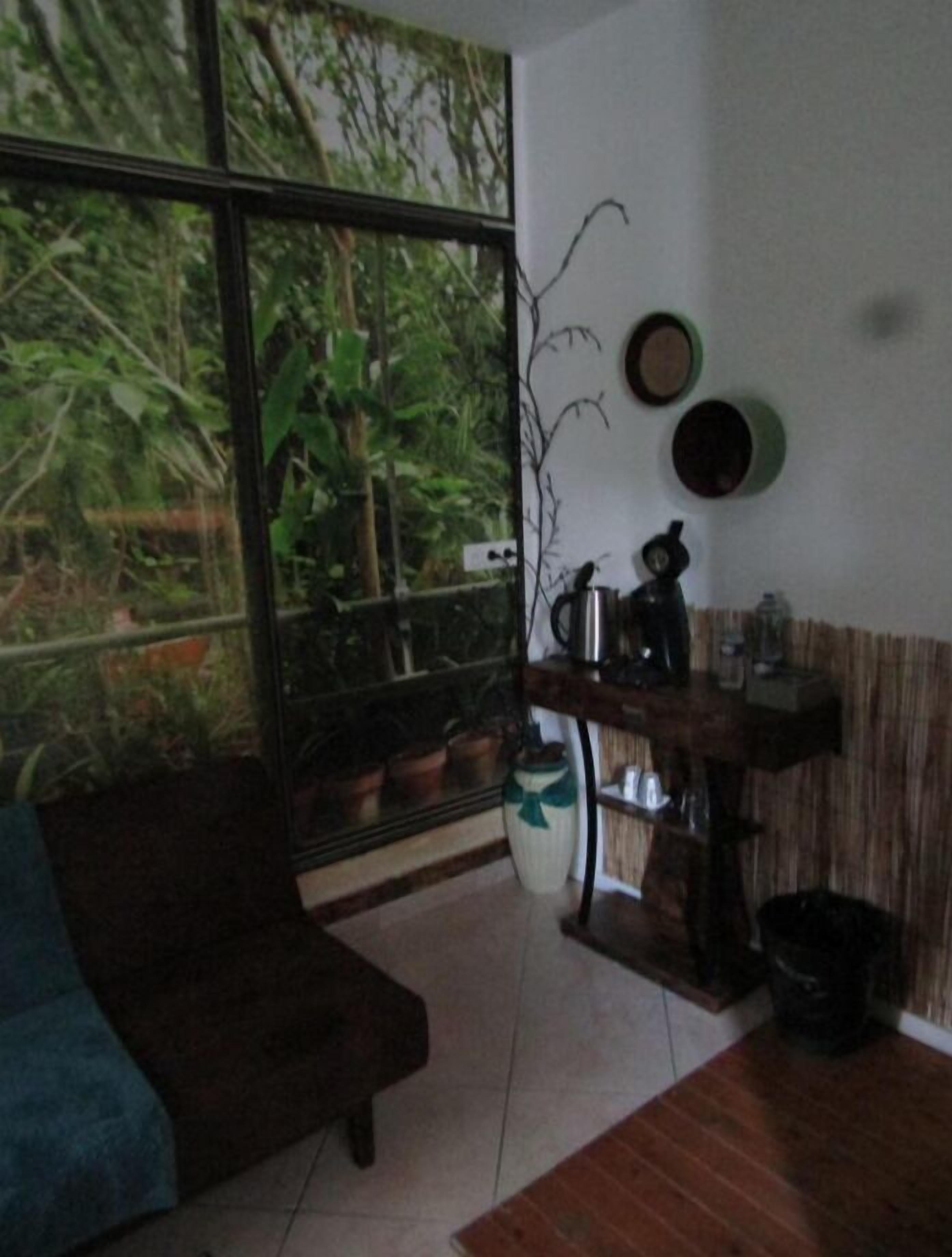 A cozy living room with a large window, a dark sofa, a coffee table, and a small wooden side table with a kettle and cups.