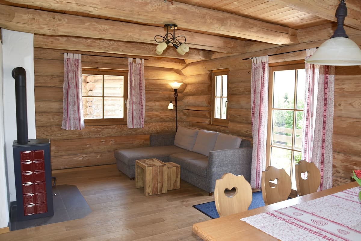 A cozy cabin interior with a wooden sofa, a dining table with chairs, a fireplace, and large windows with curtains.