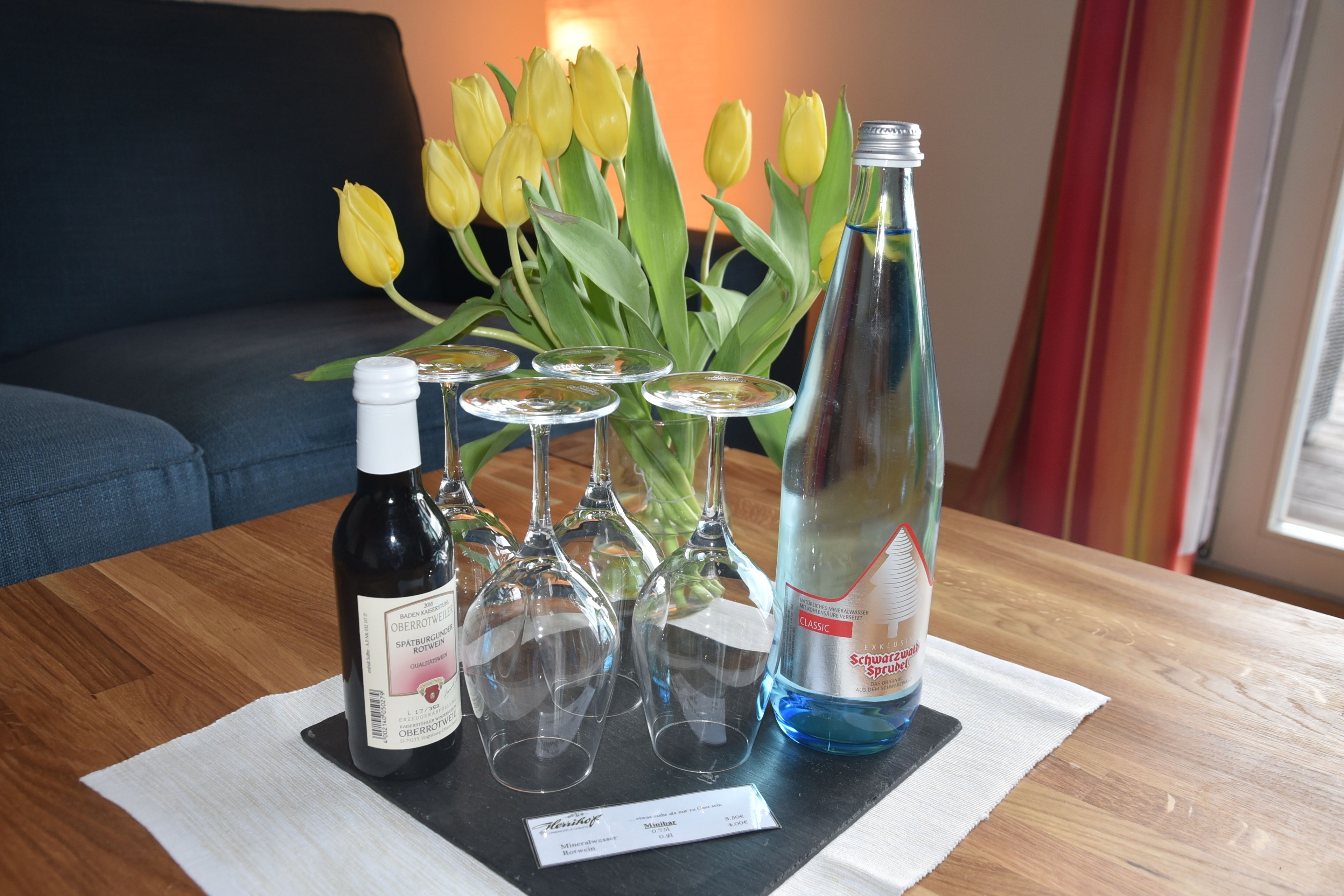 Una mesa con una botella de vino, copas de vino y una botella de agua, y un ramo de tulipanes amarillos al fondo.