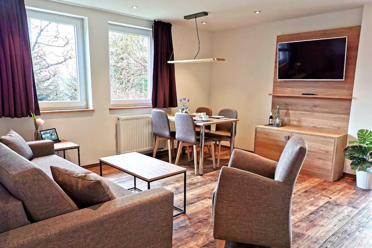 A modern living room with a sofa, armchairs, a dining table, and a TV mounted on the wall.
