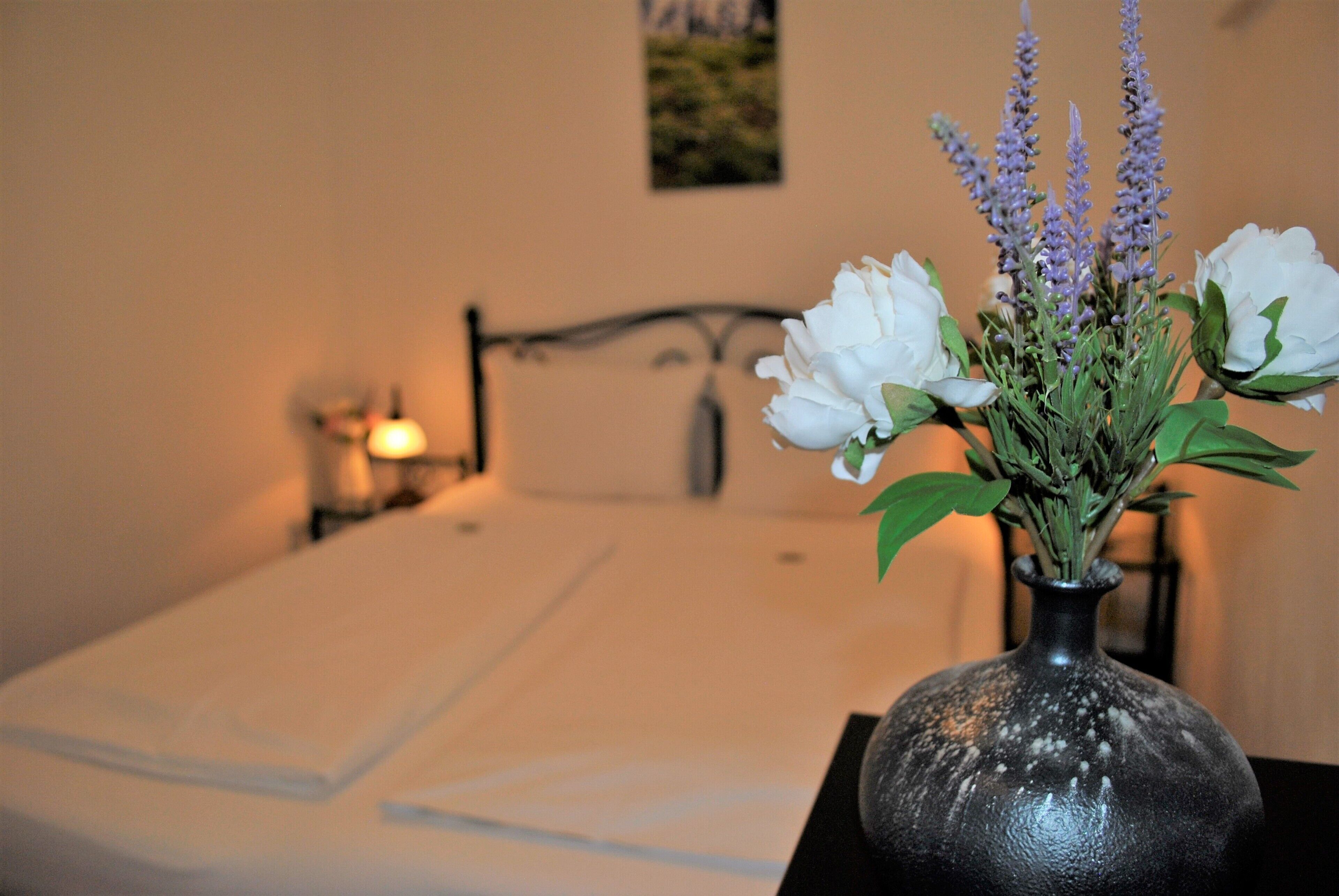 Una cama con ropa blanca, una mesita de noche con un florero y un cuadro colgado en la pared.