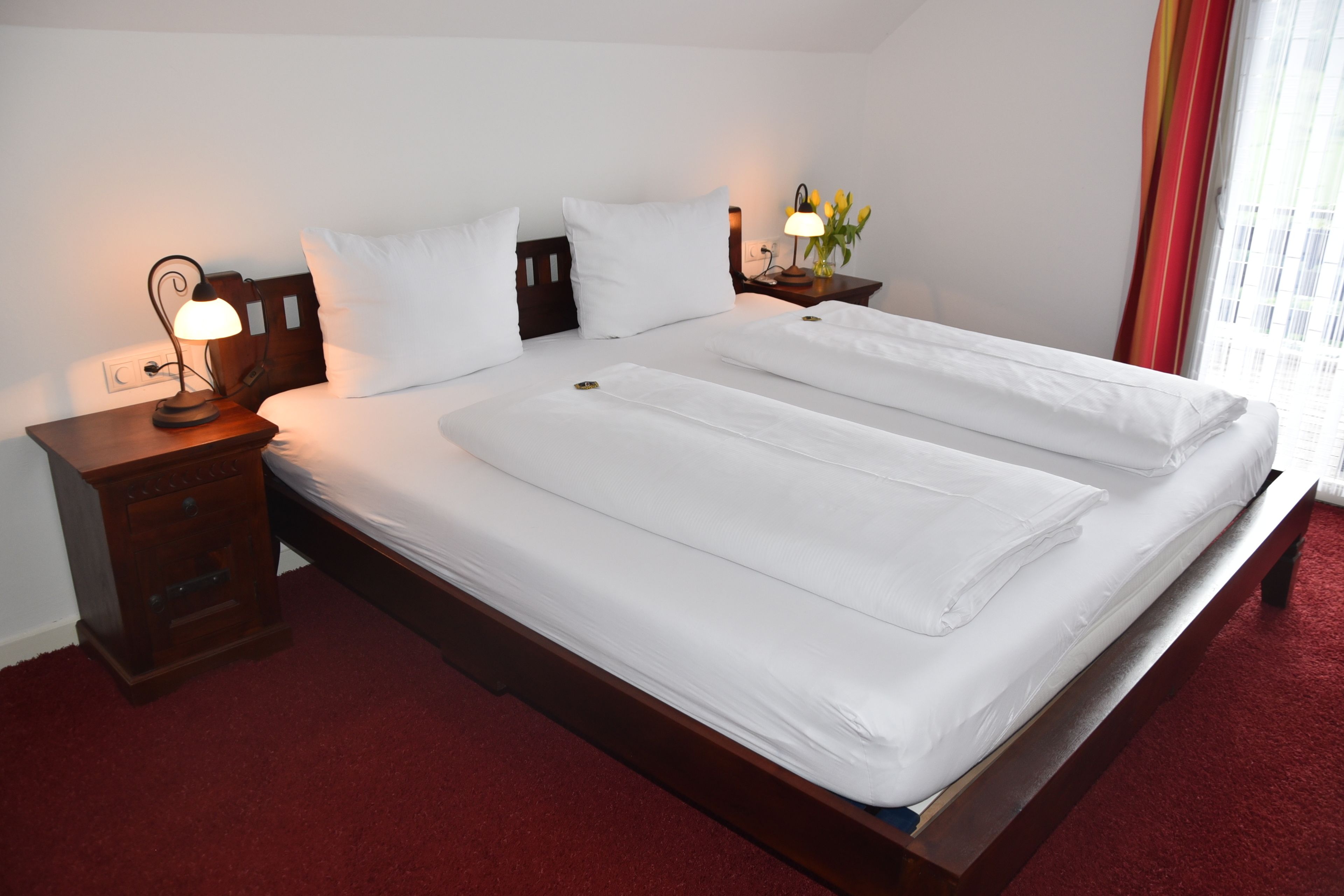 A hotel room with a large bed, wooden headboard, a nightstand with a lamp, and a window with curtains.