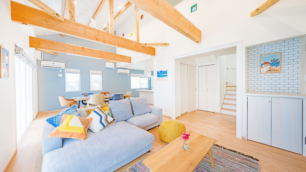 A modern interior with exposed wooden beams, a light blue sofa, a small table with a vase, and a kitchen area with a brick backsplash.