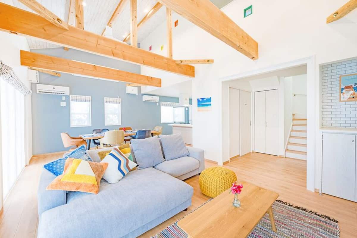 Ein modernes Interieur mit freiliegenden Holzbalken, einem hellblauen Sofa, einem kleinen Tisch mit einer Vase und einer Küchenzeile mit Ziegelrückwand.