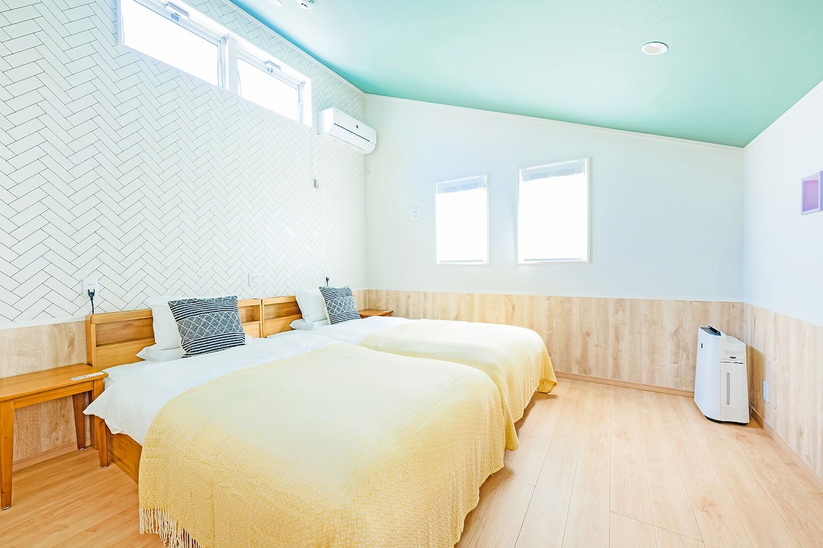 Een kamer met twee bedden, een houten bankje, een witte airconditioner en een muur met een geblokt, visgraatpatroon.
