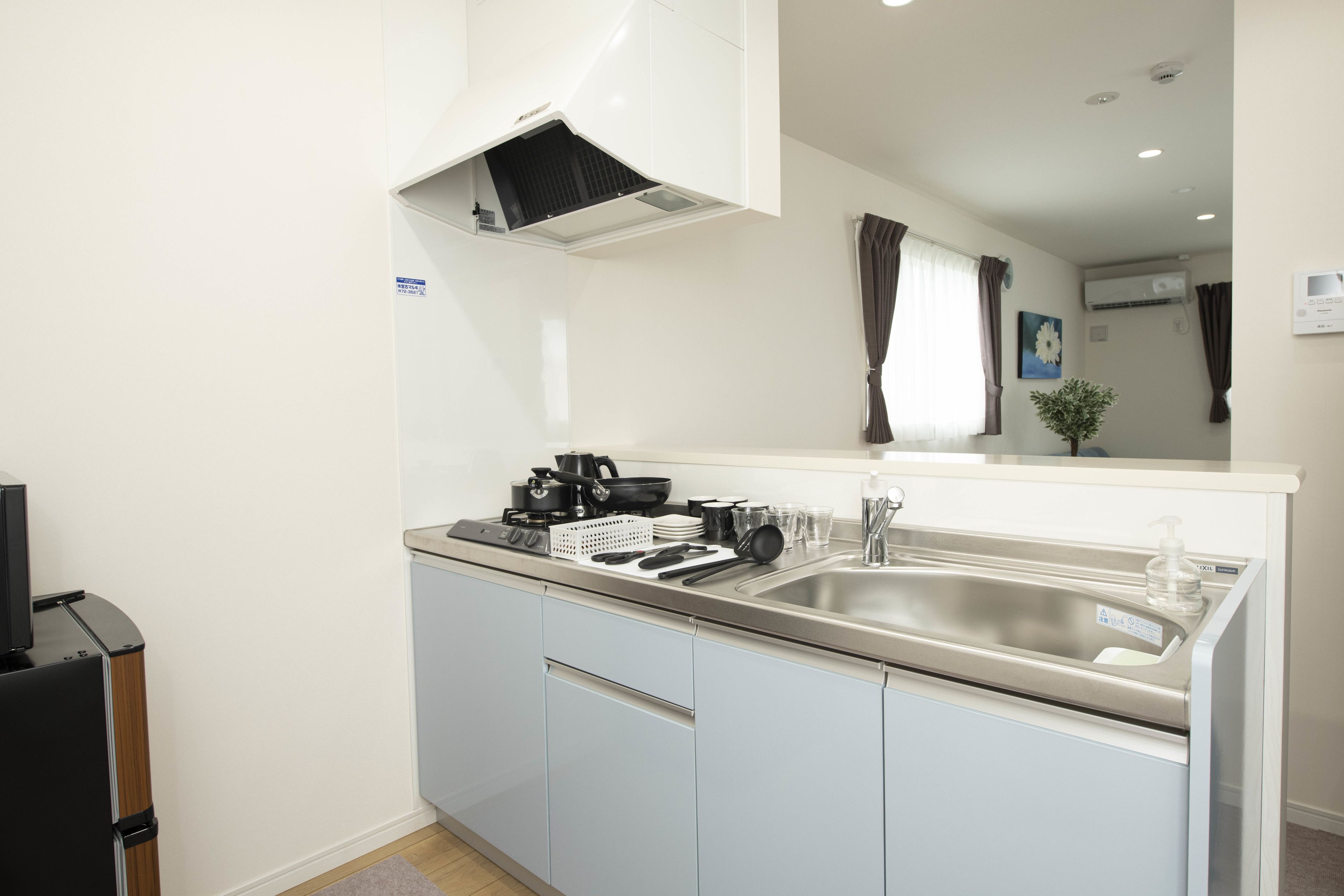 A compact kitchen with a stainless steel sink, a stove, and a microwave. There is a small countertop with a kettle and a coffee maker. A refrigerator is visible in the corner.