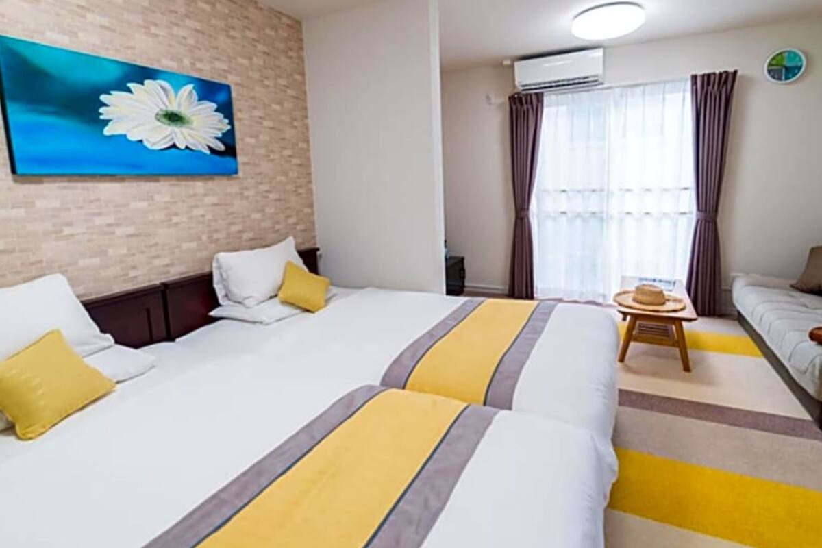 Une chambre d’hôtel avec deux lits, un canapé, une petite table sur laquelle se trouve un chapeau, et un tableau représentant une fleur accroché au mur.